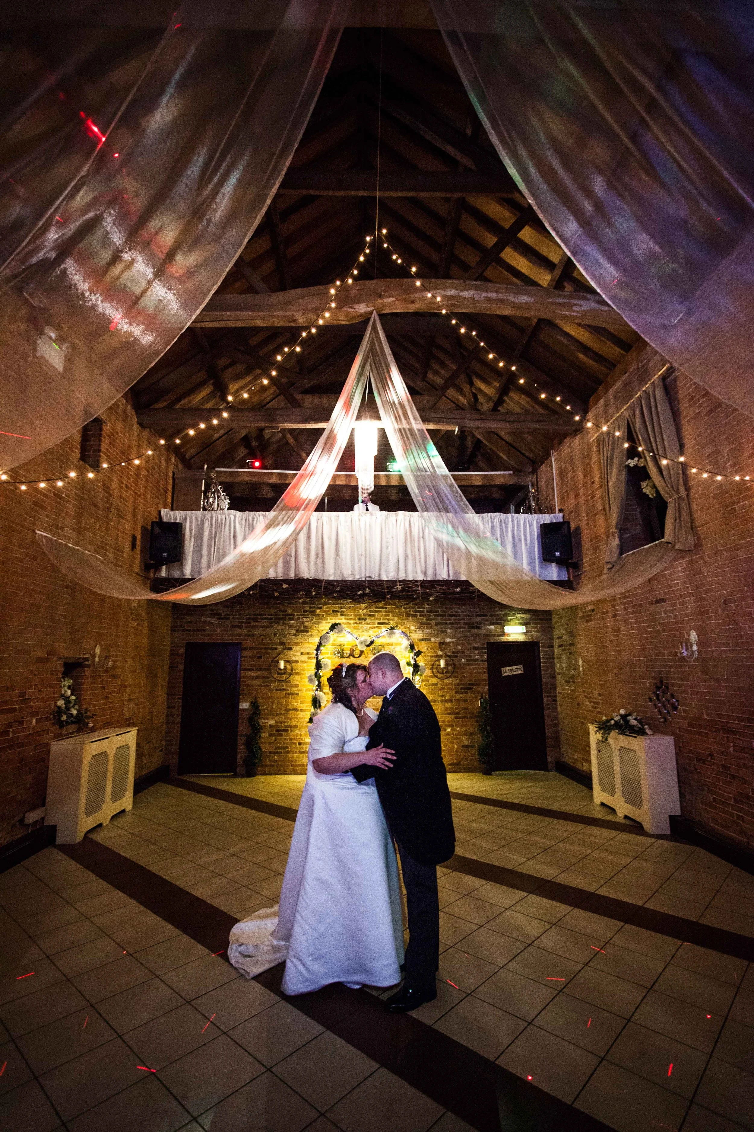 A bride and groom are sharing a kiss and dance in a warmly lit event space with brick walls, a high ceiling with exposed wooden beams, and string lights hanging overhead.