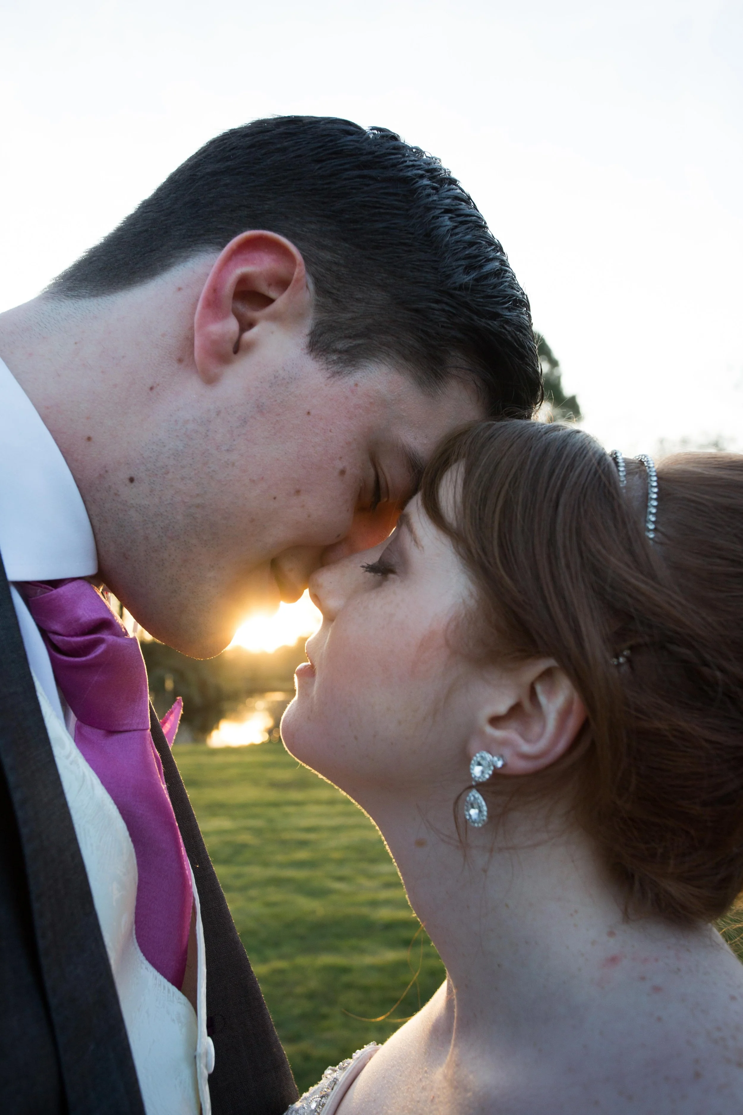 A man and woman about to kiss, with their foreheads and noses touching, outdoors during sunset; the man has dark, slicked-back hair and is wearing a tuxedo with a purple vest and bow tie, while the woman has brown hair styled with a headband and is w