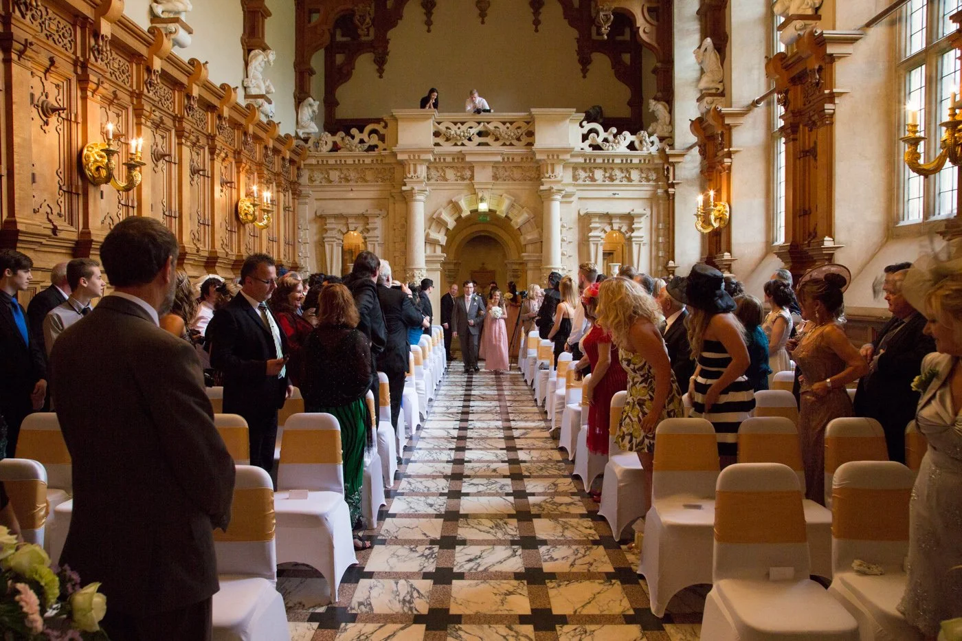 Wedding ceremony in a grand hall with guests standing along the aisle, decorative wood paneling, and large windows letting in natural light.