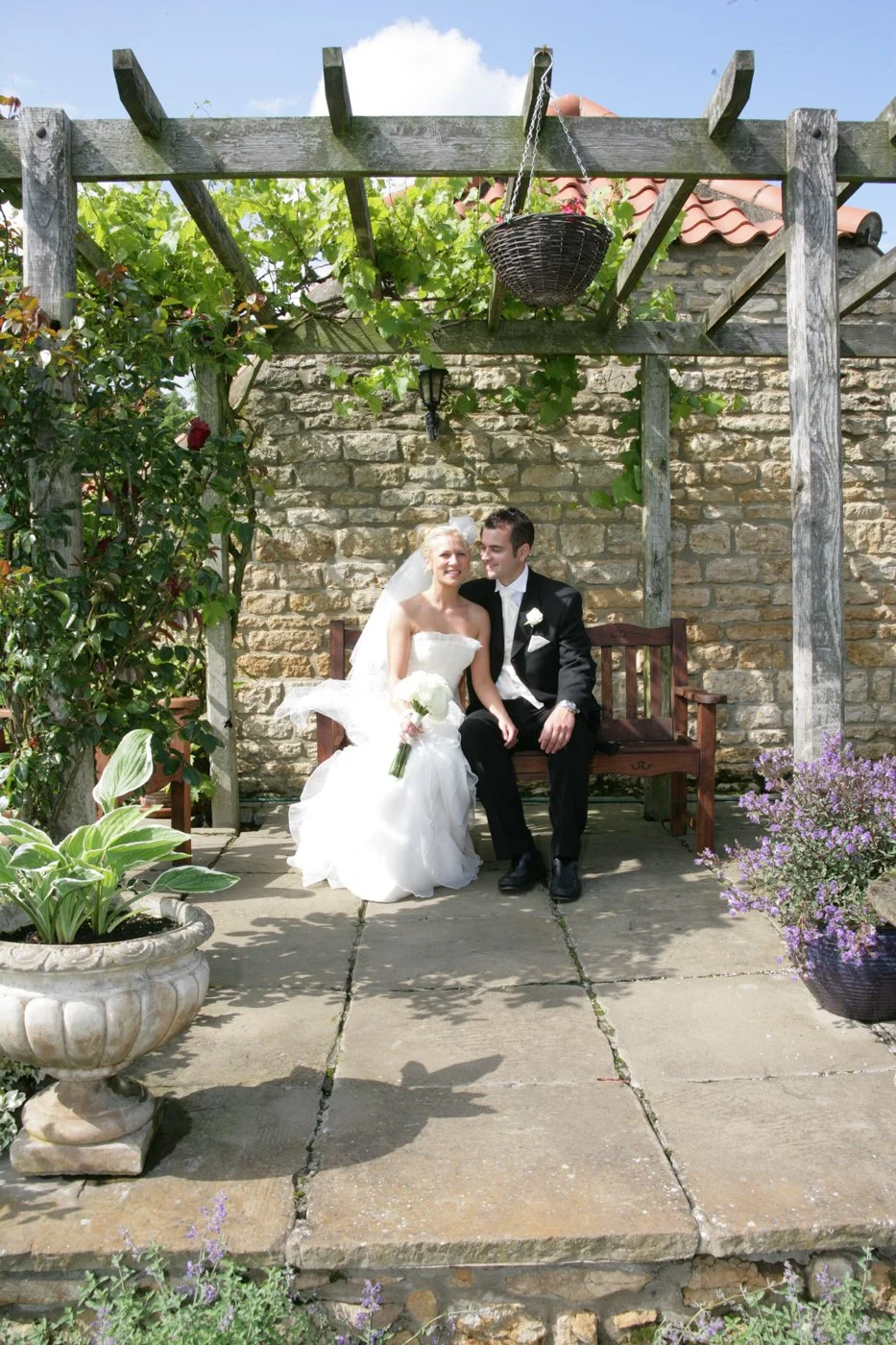 A bride and groom sitting on a wooden bench outdoors, surrounded by plants and flowers, with a stone wall and pergola overhead.