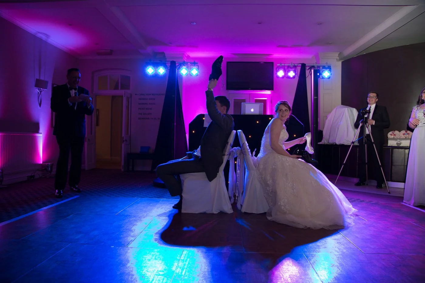 Bride and groom at their wedding reception, seated back to back on chairs, with the groom raising his leg and the bride smiling, under colorful wedding lighting.