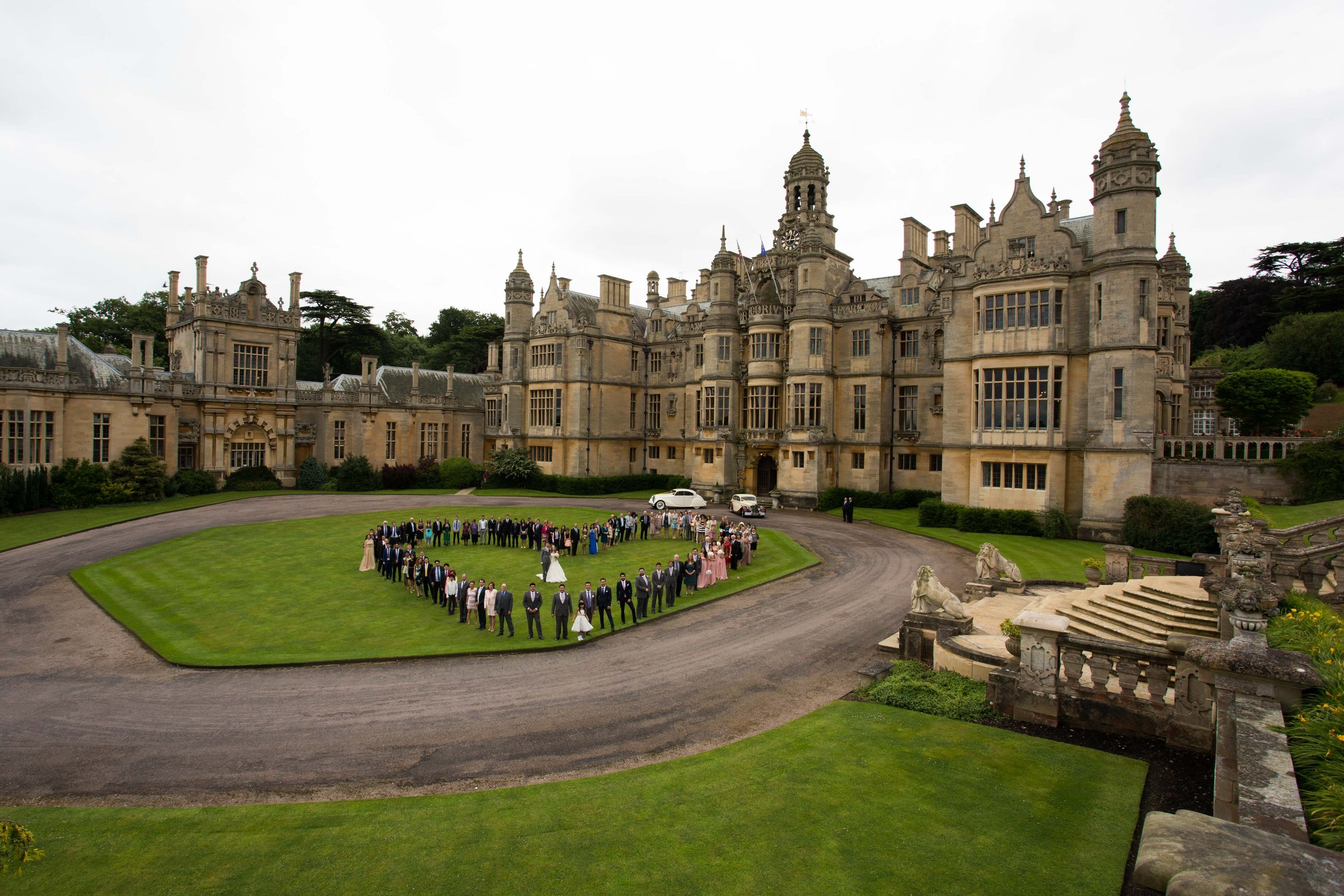 A large group of people, including a bride in a white gown and a groom in a black tuxedo, poses on the lawn of a historic castle during a wedding.