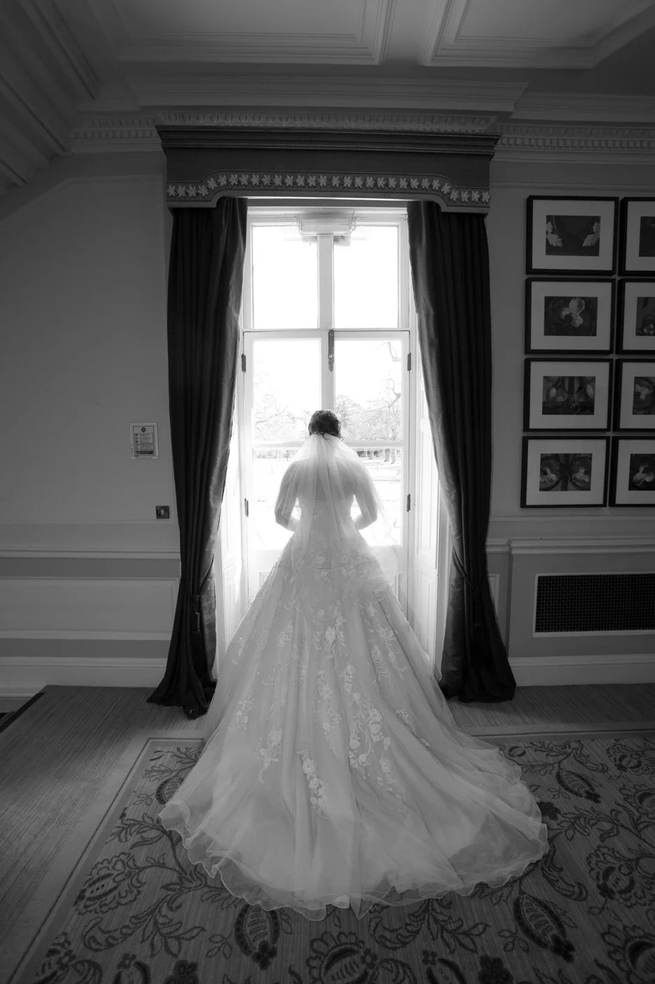 A bride in a wedding gown standing in front of a large window in an elegant room, gazing outside.