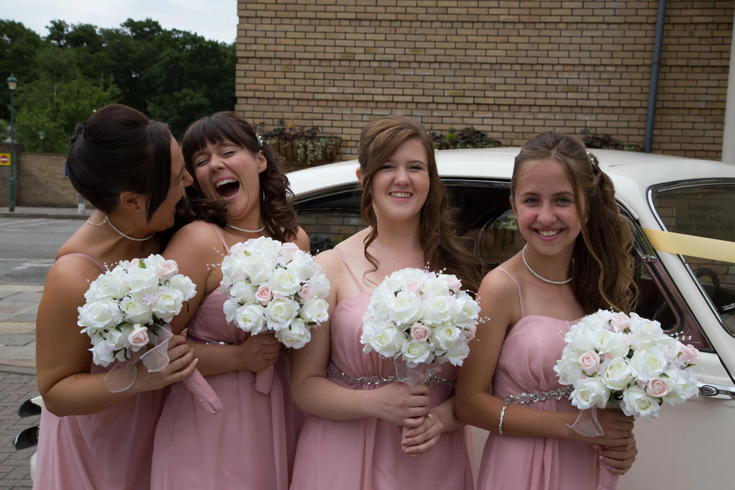 Four women in pink dresses holding white and pale pink bouquets standing in front of a yellowish classic car, with a brick wall and green trees in the background.