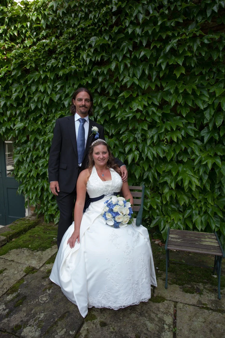 A bride and groom pose for a wedding photo outdoors in front of a lush green ivy-covered wall, with the bride sitting on a chair holding a bouquet of blue and white flowers, and the groom standing behind her with his arm around her.