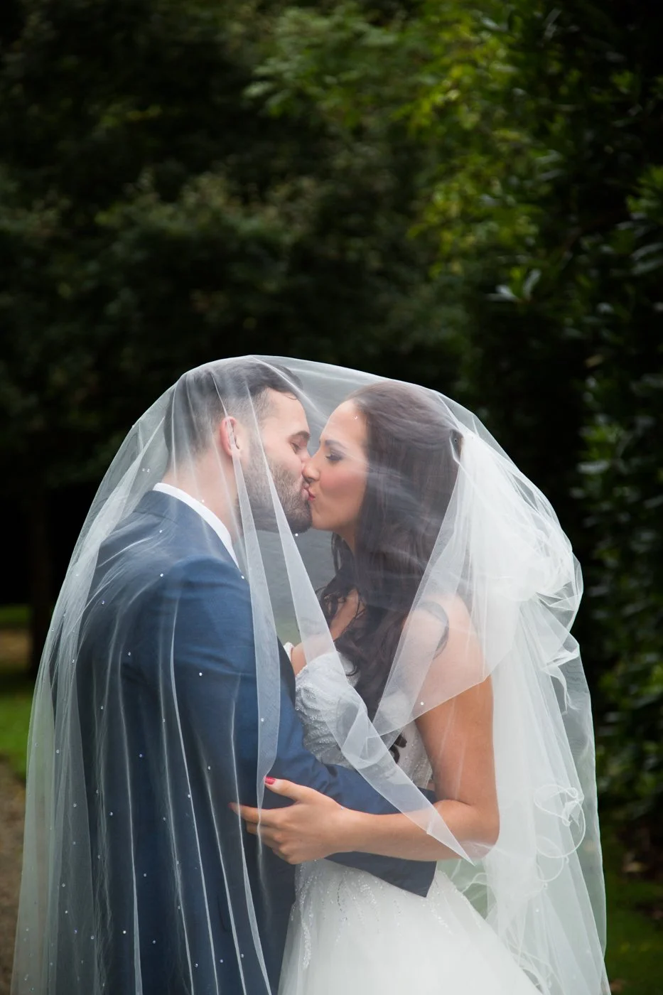 A bride and groom sharing a kiss under a veil in an outdoor setting with green trees in the background.