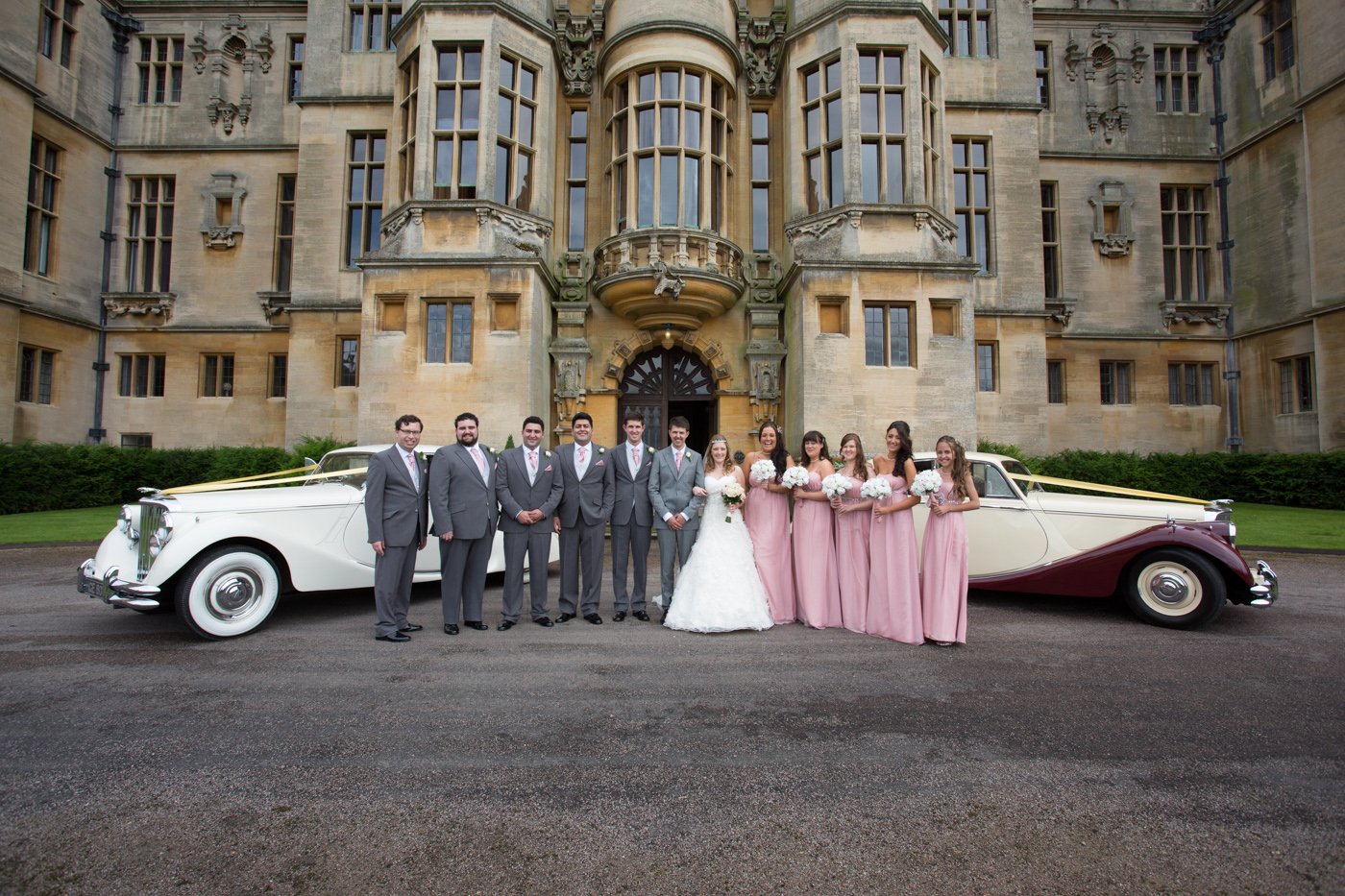 A wedding party standing in front of a historic castle, with a vintage white and maroon car decorated with ribbons parked behind them. The group includes men in gray suits, women in pink dresses holding bouquets, and a bride in a white wedding gown holding a bouquet.