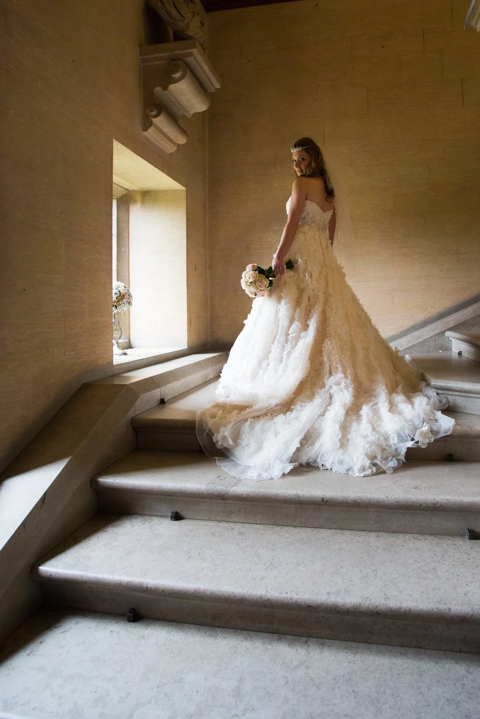 A bride in a white wedding gown standing on a staircase with a bouquet of flowers, near a window with natural light illuminating her and the wall behind her.