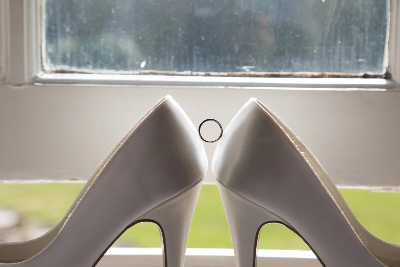 A pair of white high-heeled shoes placed on a windowsill, with the heels touching, and a ring between them, sunlight coming through the window.
