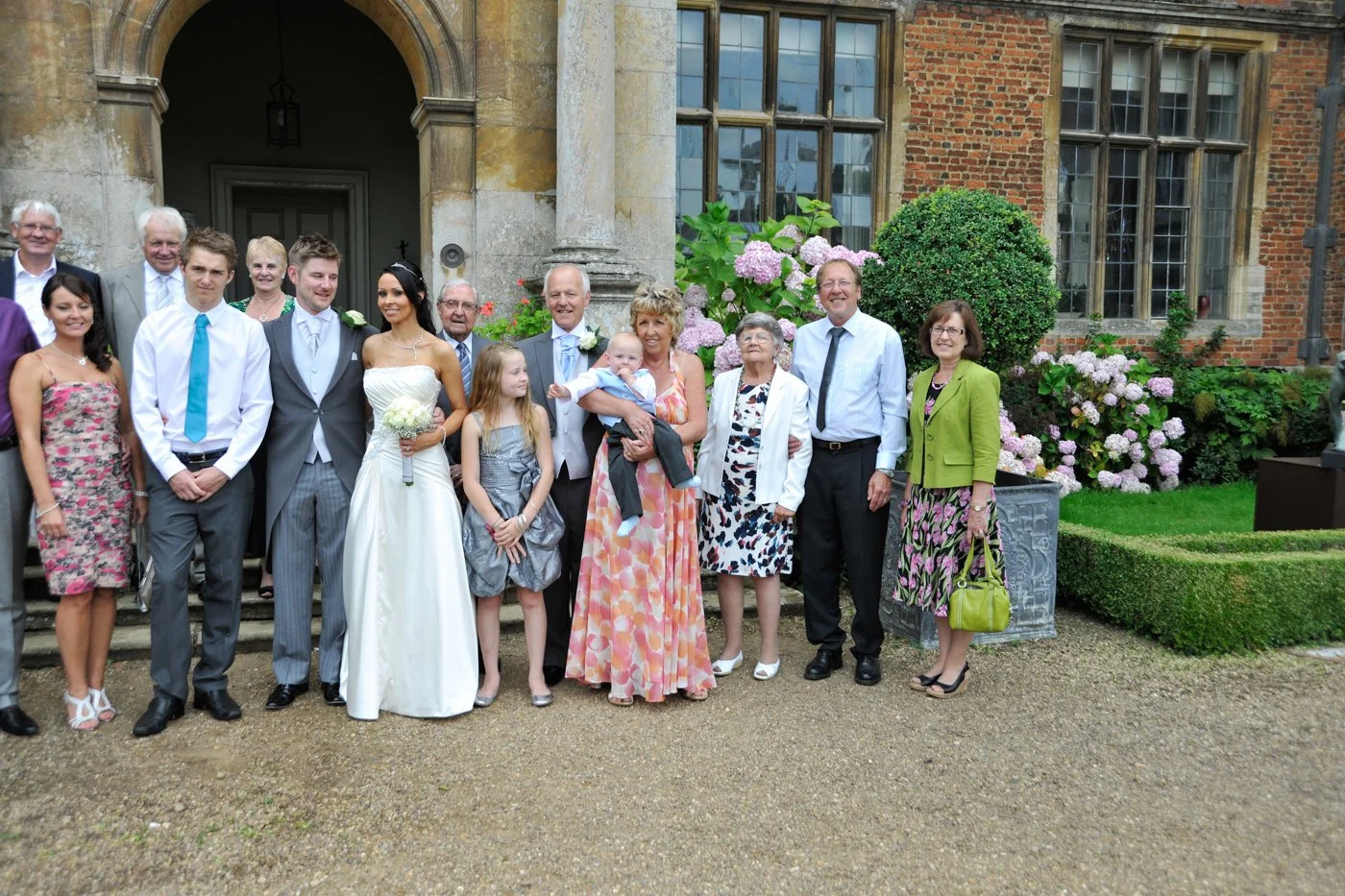 Group of people in wedding attire posing outside a historic brick building with hydrangeas and greenery.