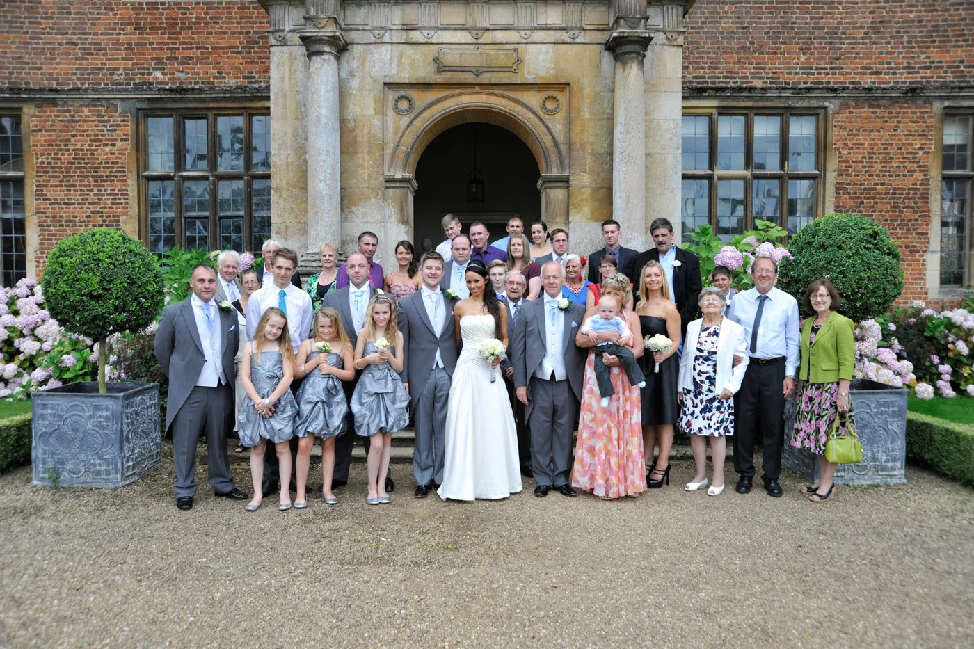 A large group of wedding guests and family members posing for a photo outside a historic brick building with lush greenery and flower arrangements.