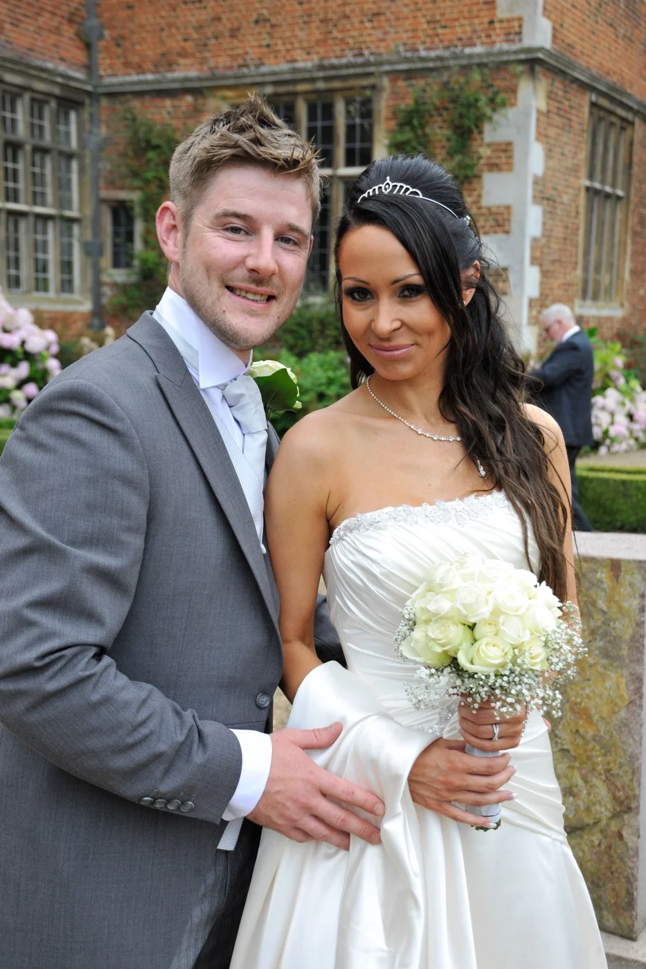 Bride and groom at a wedding outdoors, with a brick building and garden in the background, smiling and posing for the photo.