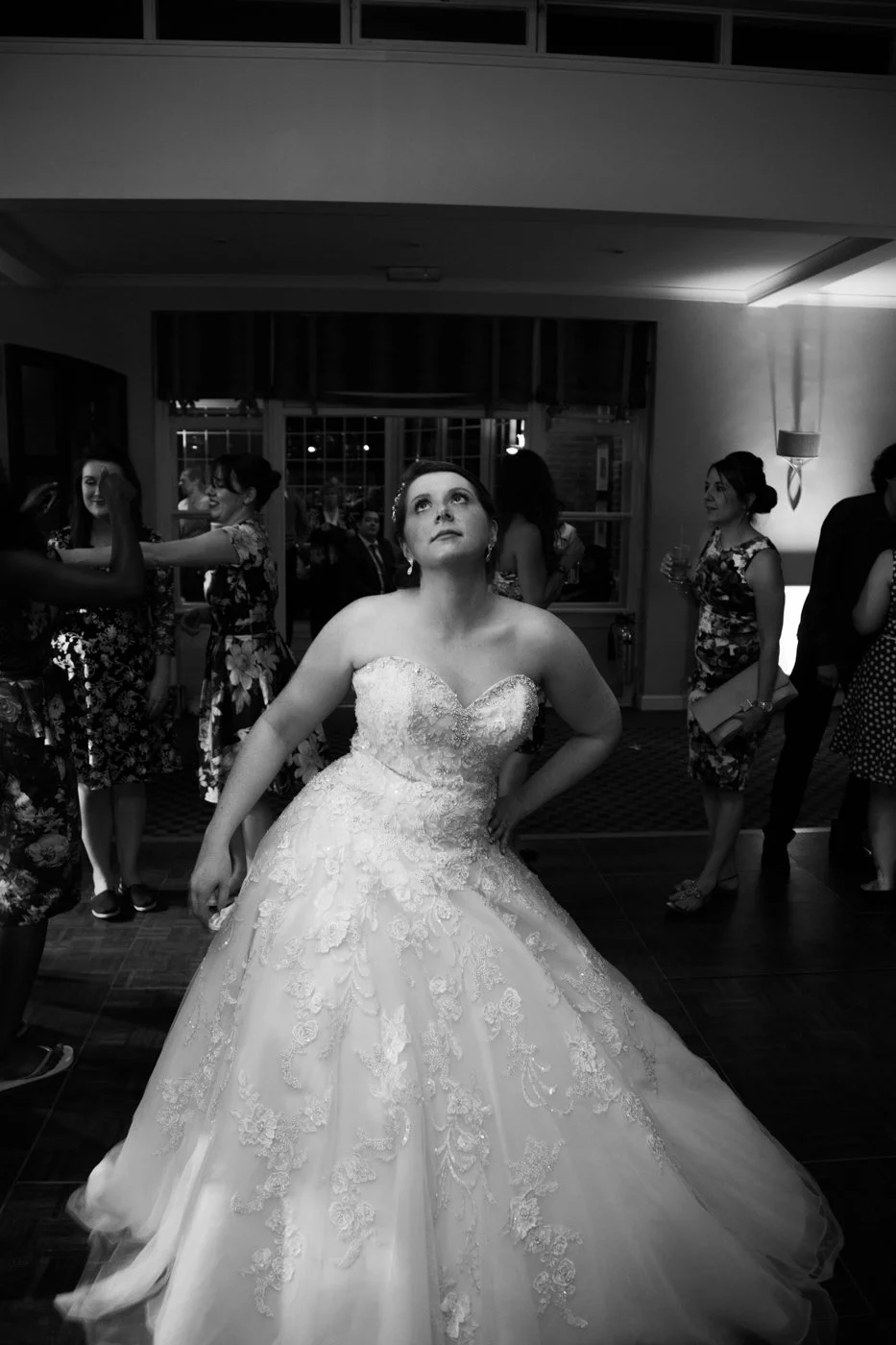 Black and white photo of a bride in a wedding dress at a reception, surrounded by guests in a decorated indoor venue.