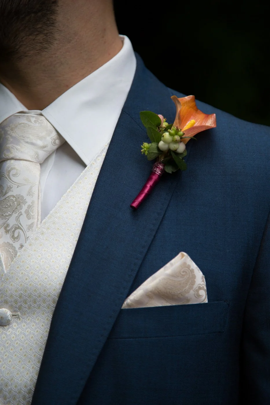 Close-up of a groom's navy blue suit with a floral boutonniere on the lapel, featuring a peach-colored flower, green leaves, white berries, and a deep pink wrap. He is wearing a white dress shirt, an ornate cream-colored vest, a matching cream-colore