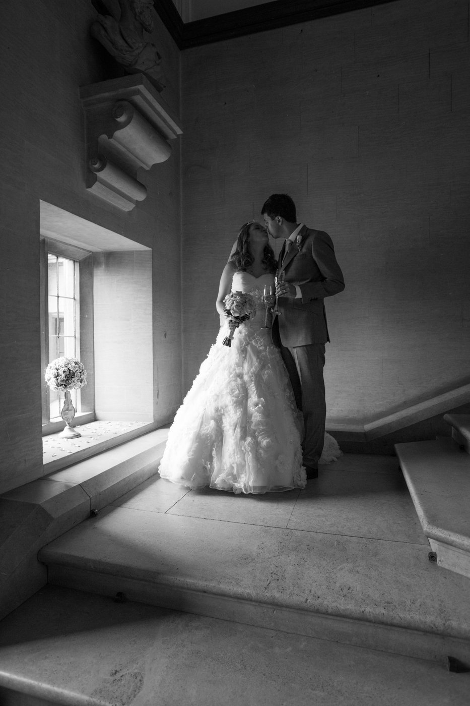 A black and white photograph of a bride and groom standing close and kissing on a staircase inside a building, with the bride holding a bouquet and the groom holding a glass of champagne.