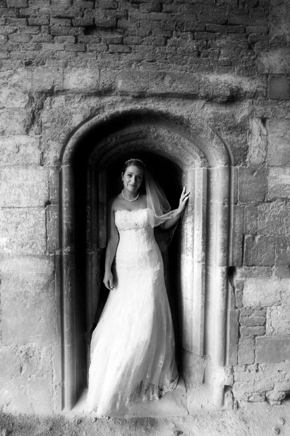 Black and white photo of a bride in a wedding dress standing in a stone archway, smiling and holding the edge of the arch.