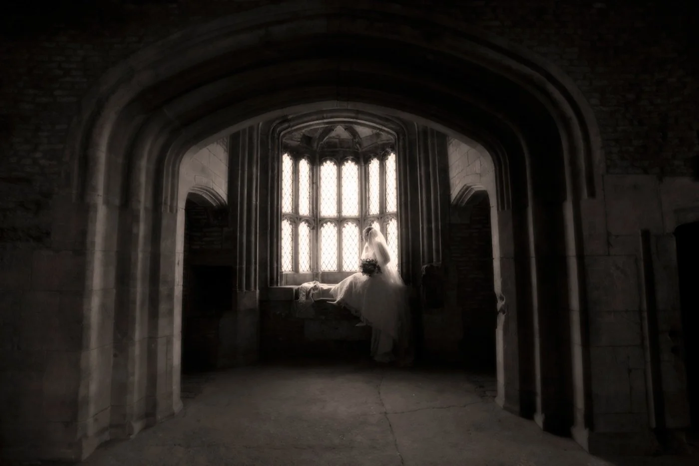 A bride in a wedding dress looking out a large arched stained glass window in a historic stone building.