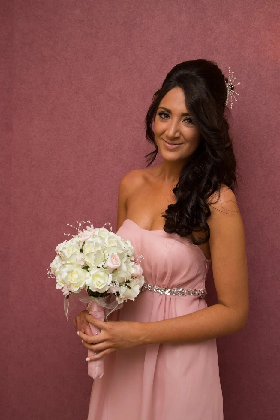 A woman in a pink strapless dress holding a bouquet of white and pink roses stands against a pink background, smiling.