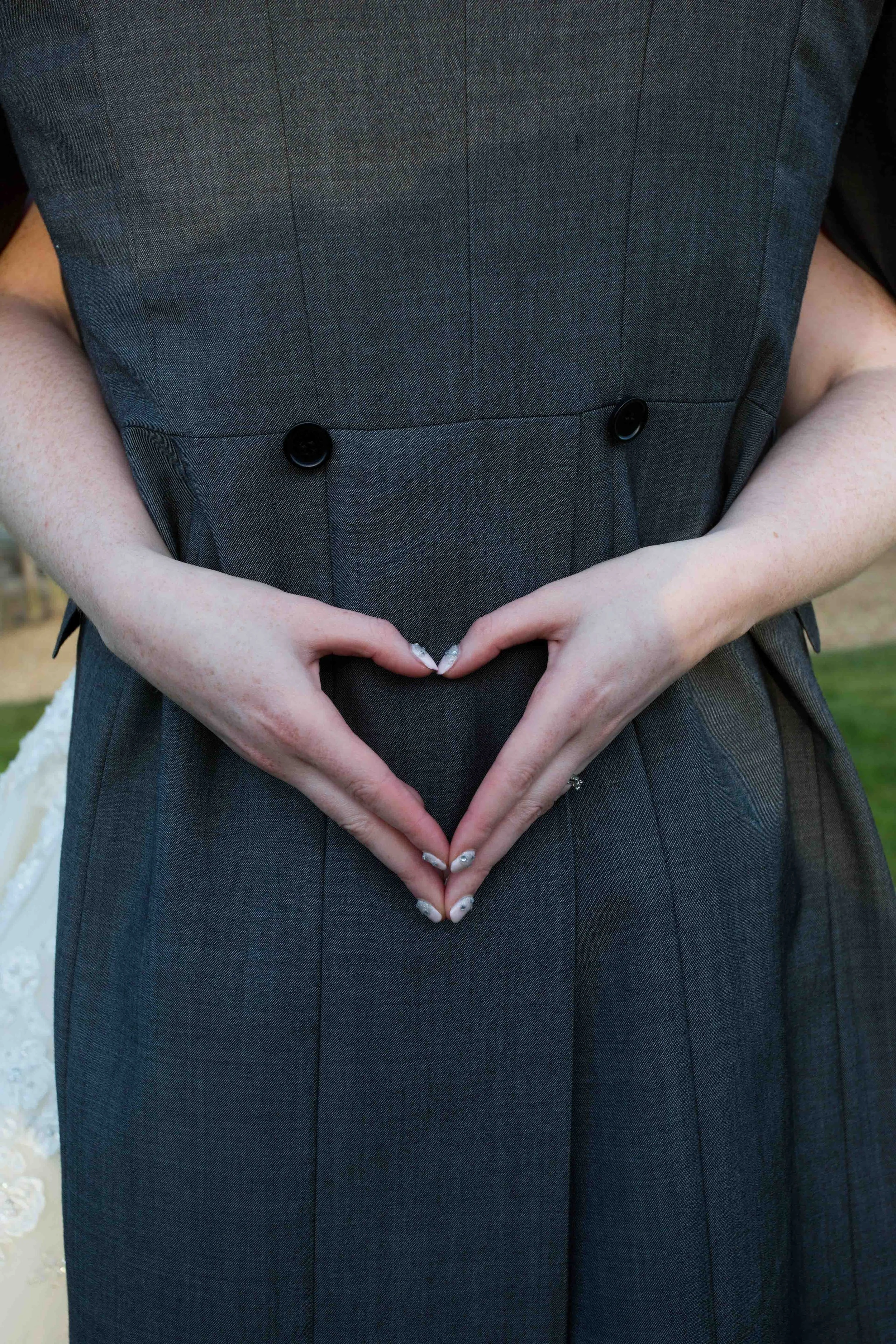 Close-up of a person wearing a dark gray, pinstripe dress or suit, making a heart shape with their hands. The person's arms are visible, and the background is blurry.