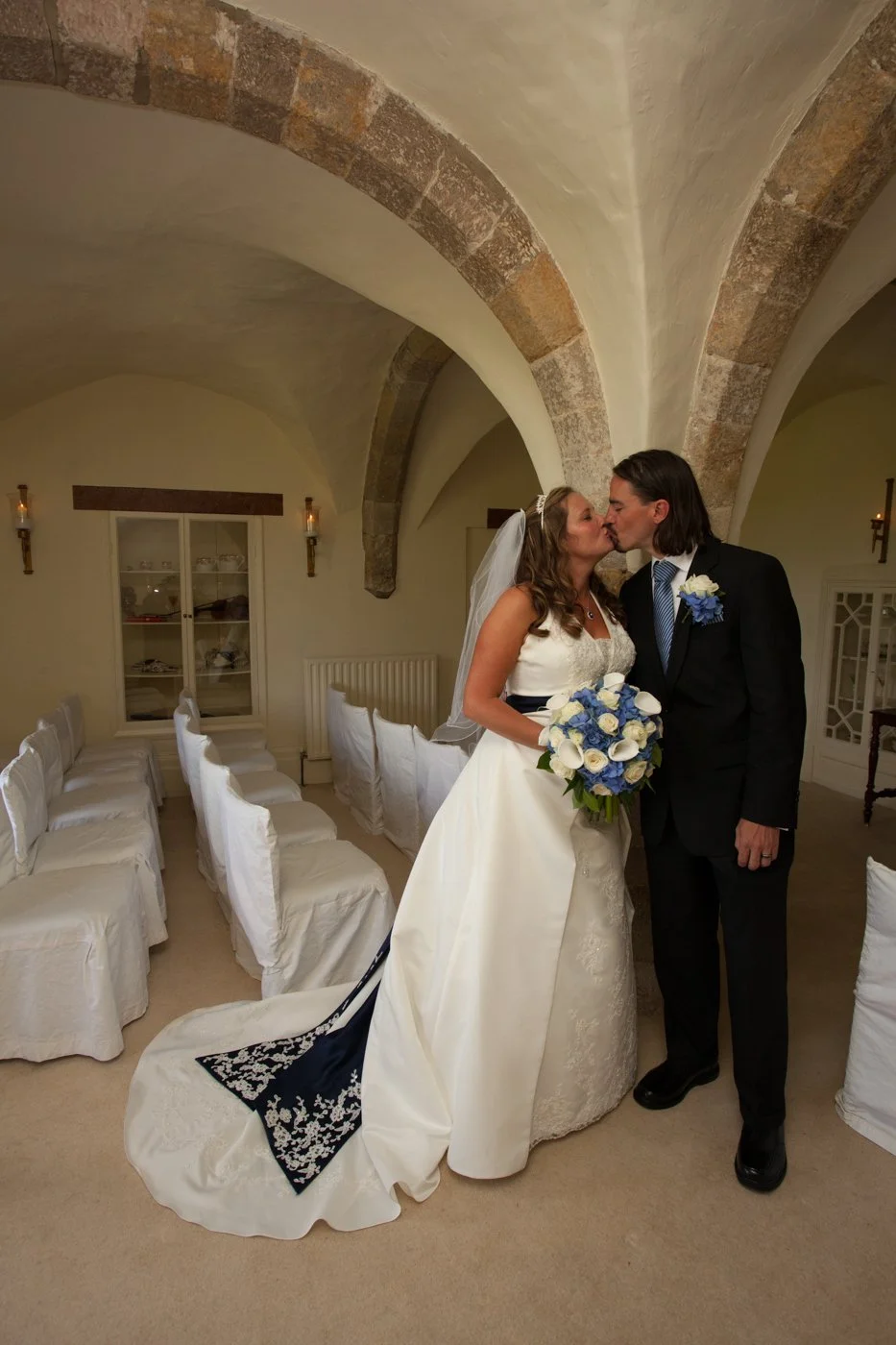 A bride and groom kissing in a room with arched stone ceiling, white chairs covered with white cloth, and a cabinet in the background.
