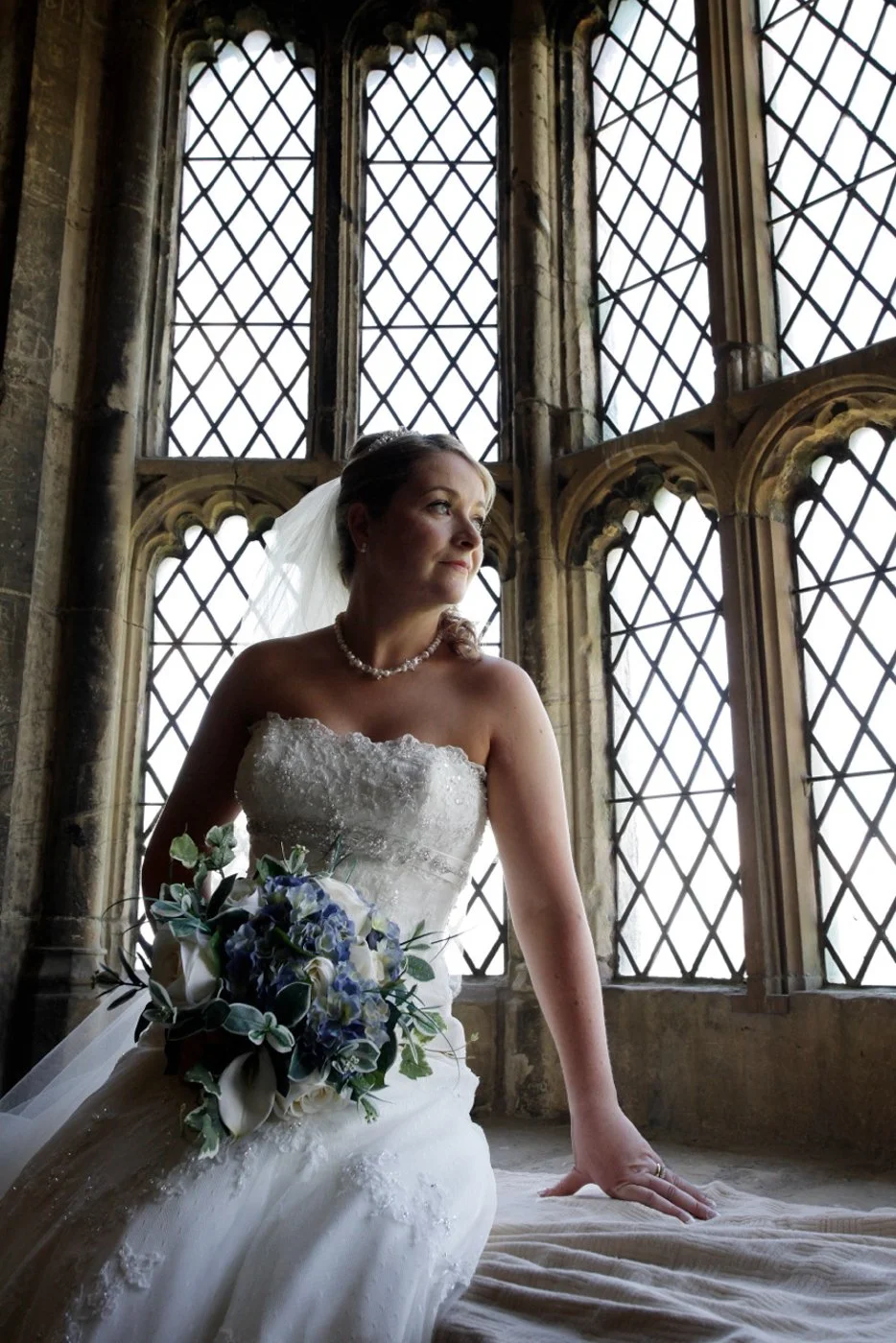 A bride in a strapless wedding gown holding a bouquet of blue and white flowers, sitting by a large window with ornate stone framing, gazing out.