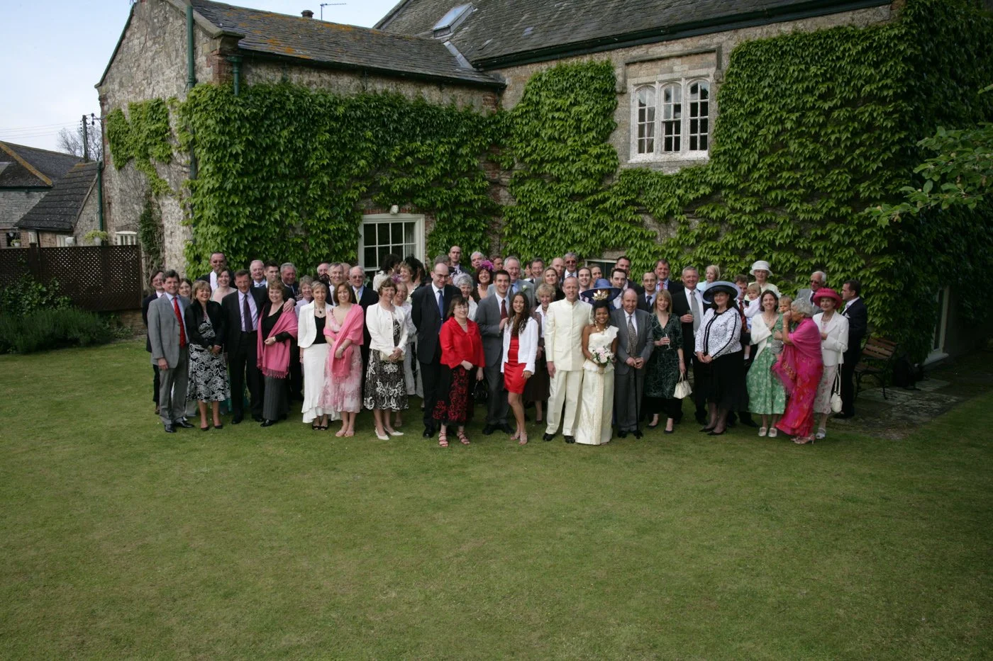 Group of people gathered outside a stone building covered in green ivy, celebrating a wedding. The bride and groom are in the center, with guests dressed in formal attire.