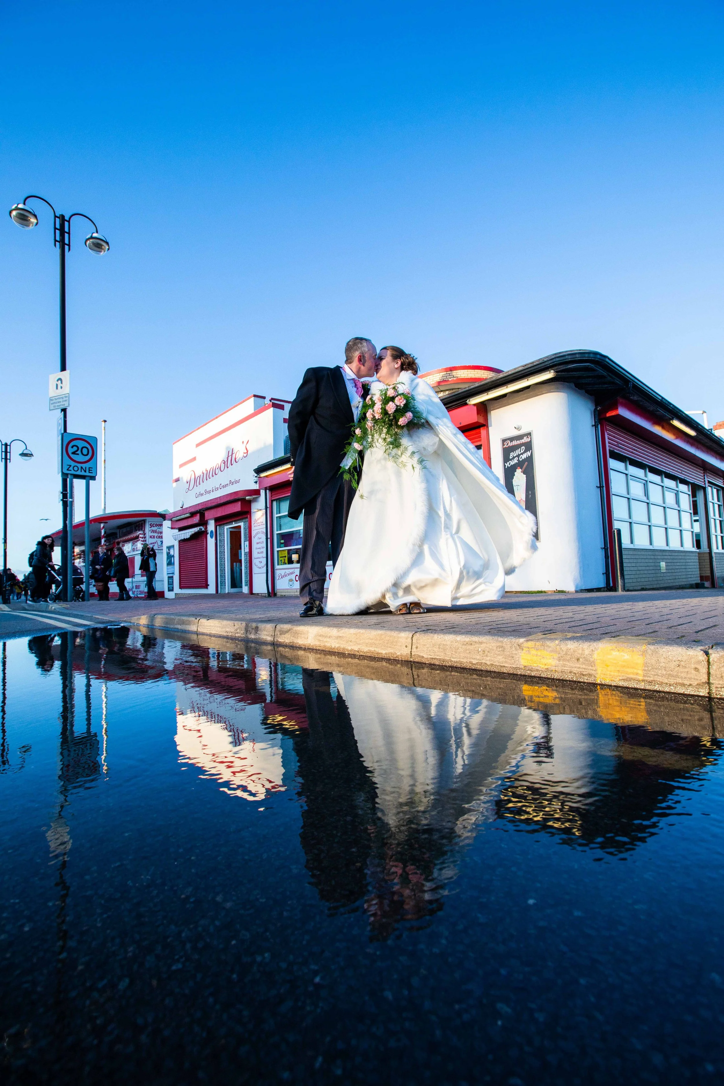 A bride and groom share a kiss on a street corner, with the bride holding a bouquet of pink and white flowers. The reflection is seen in a puddle on the pavement, with a blue sky overhead and a red and white building in the background.
