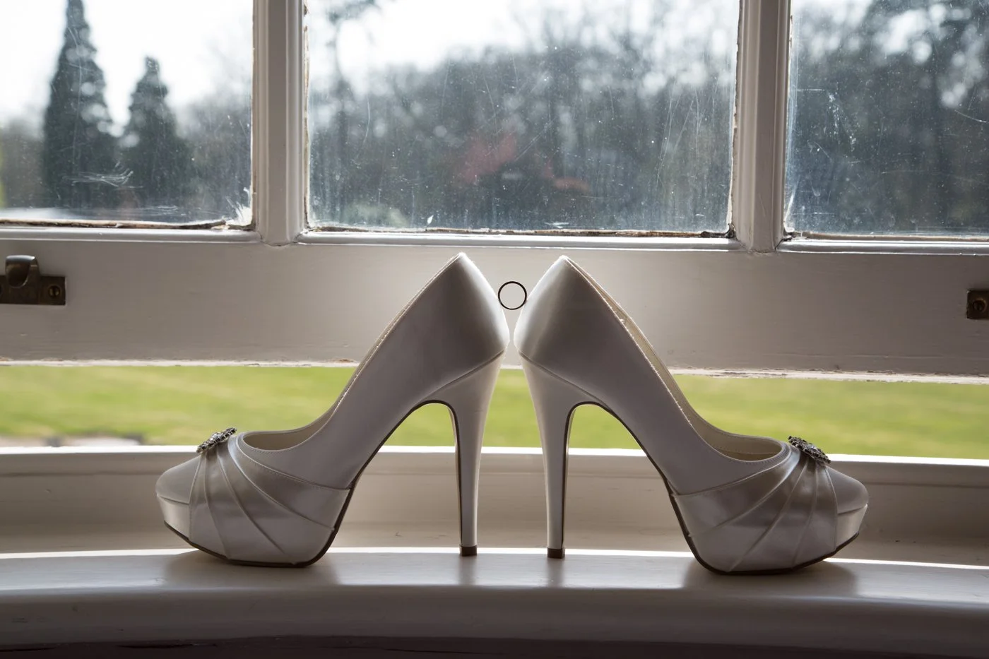 A pair of white high-heeled wedding shoes with decorative bows on the toe, placed on a windowsill with a view of blurred green grass and trees outside.