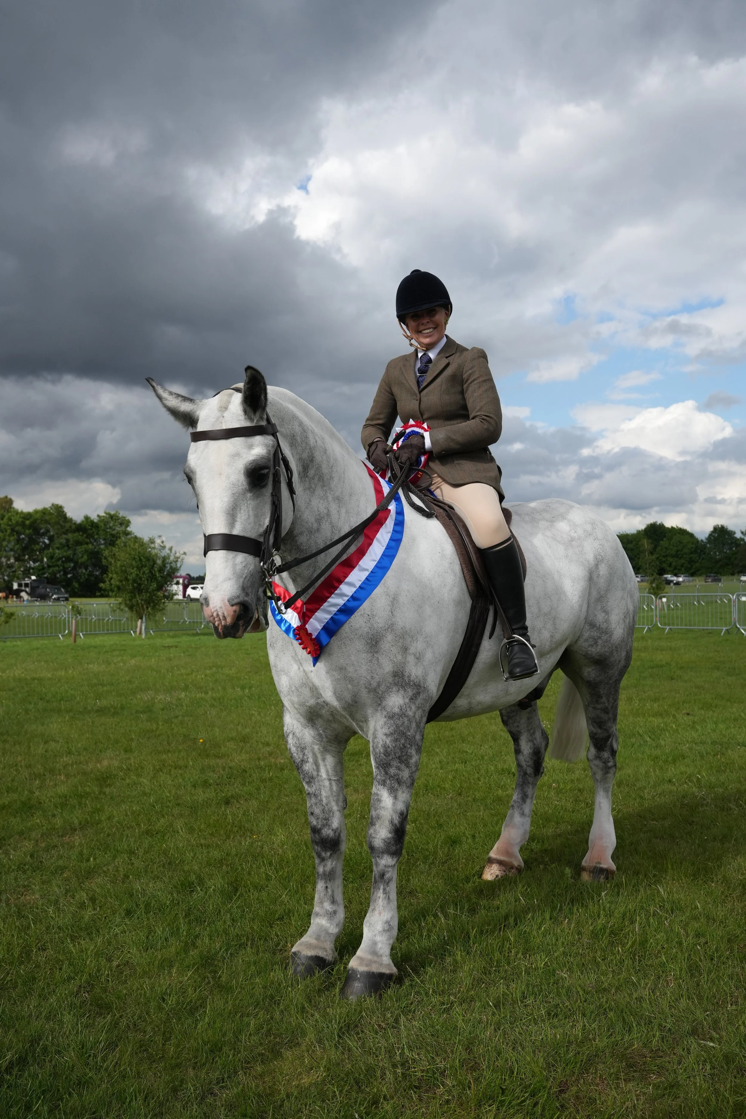 A woman dressed in equestrian attire, including a helmet, blazer, and riding pants, sitting atop a gray horse with a decorated ribbon on its neck in an outdoor grassy area during a cloudy day, indicating a competition or event.