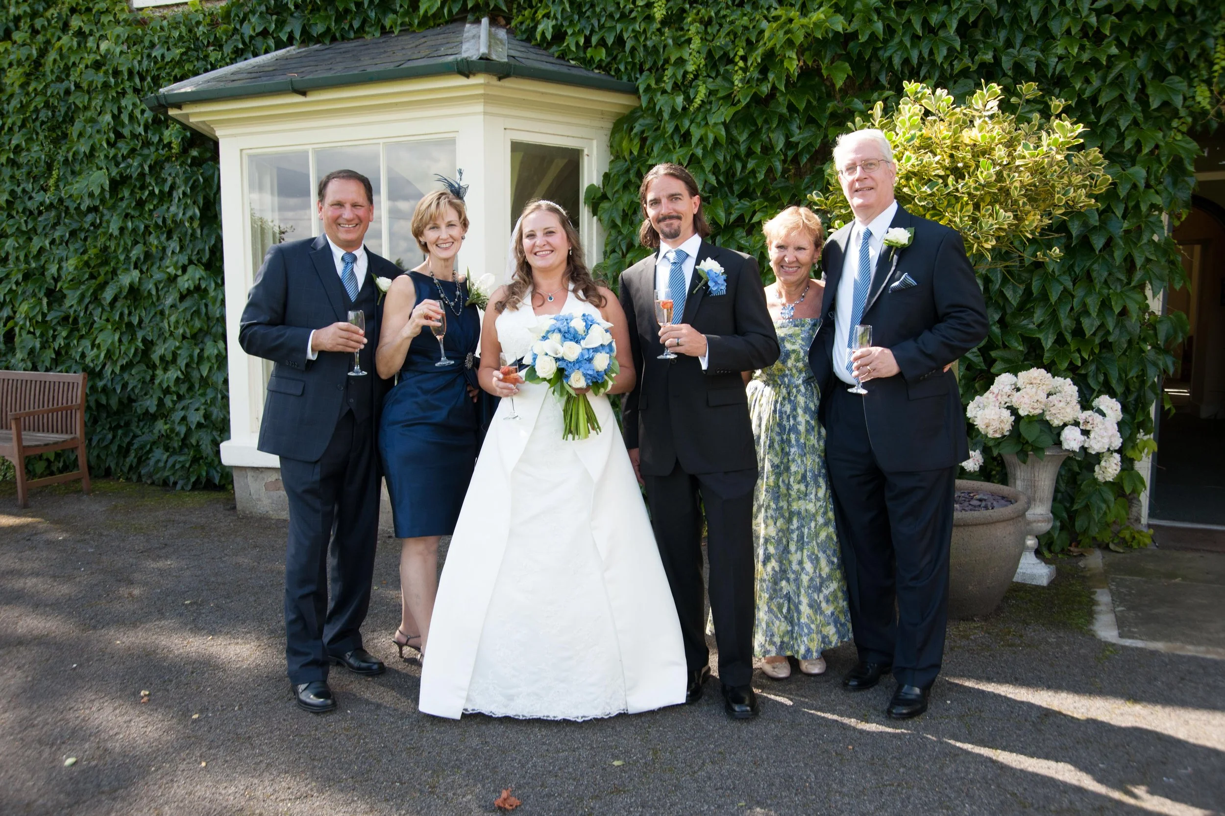Group of six people at a wedding, standing in front of a ivy-covered building, holding champagne glasses, with the bride holding a bouquet of white and blue flowers.