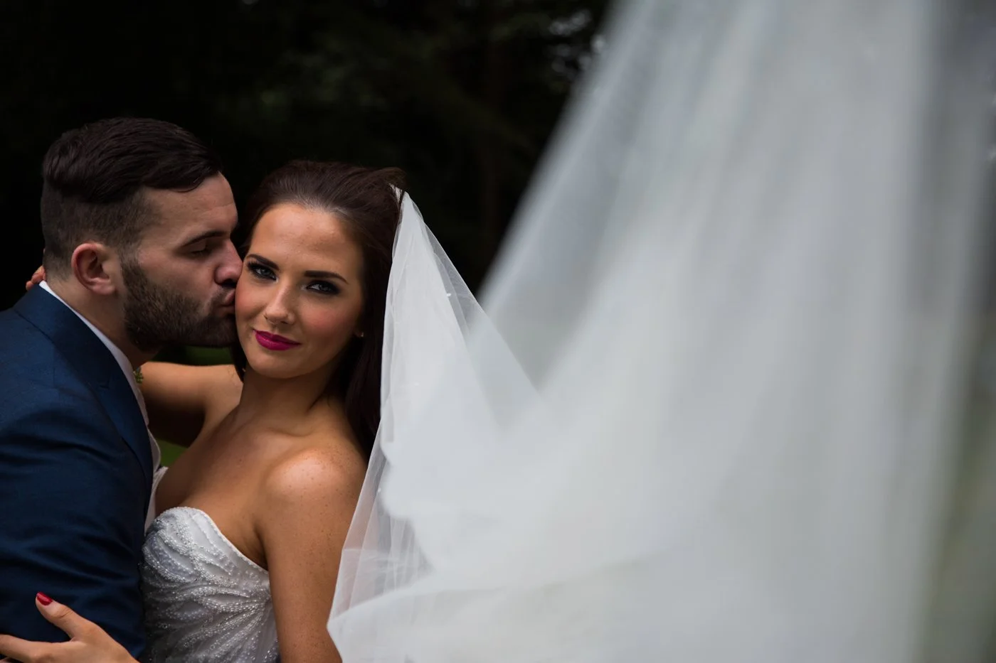 A newlywed couple outdoors, with the groom kissing the bride's cheek as she looks at the camera, wearing a white wedding dress and veil.