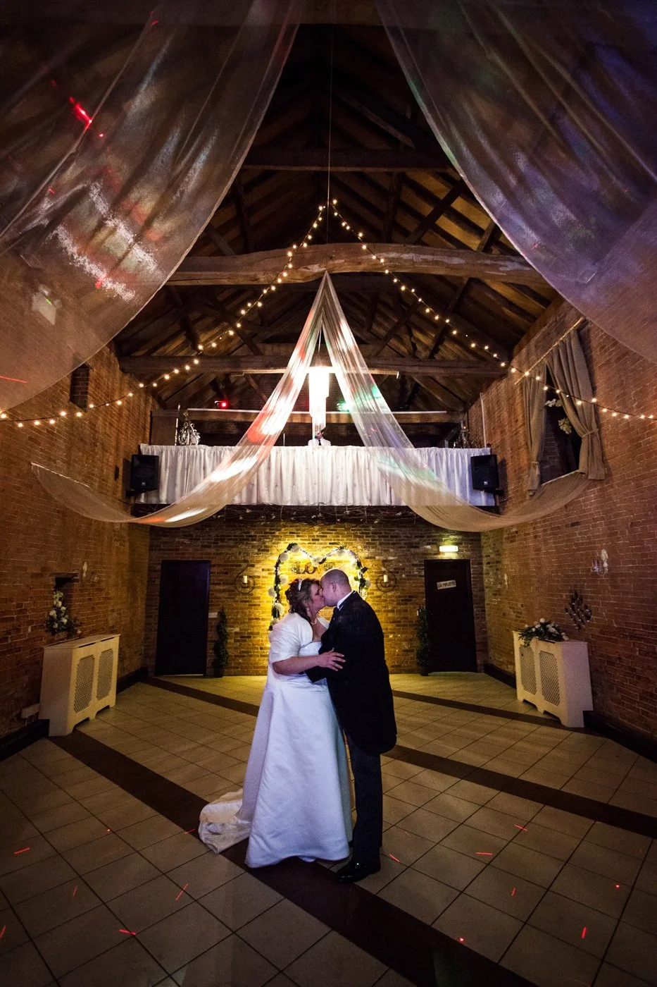 Couple in wedding attire sharing a kiss on the dance floor of a decorated rustic wedding venue with string lights and draped fabric overhead.