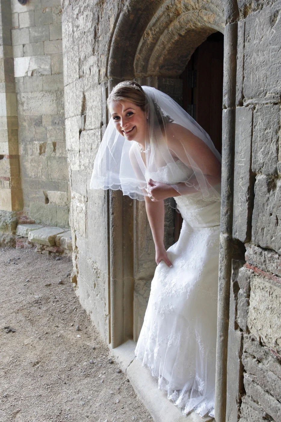 A bride with a veil and wedding dress leaning out of a small stone window, smiling.