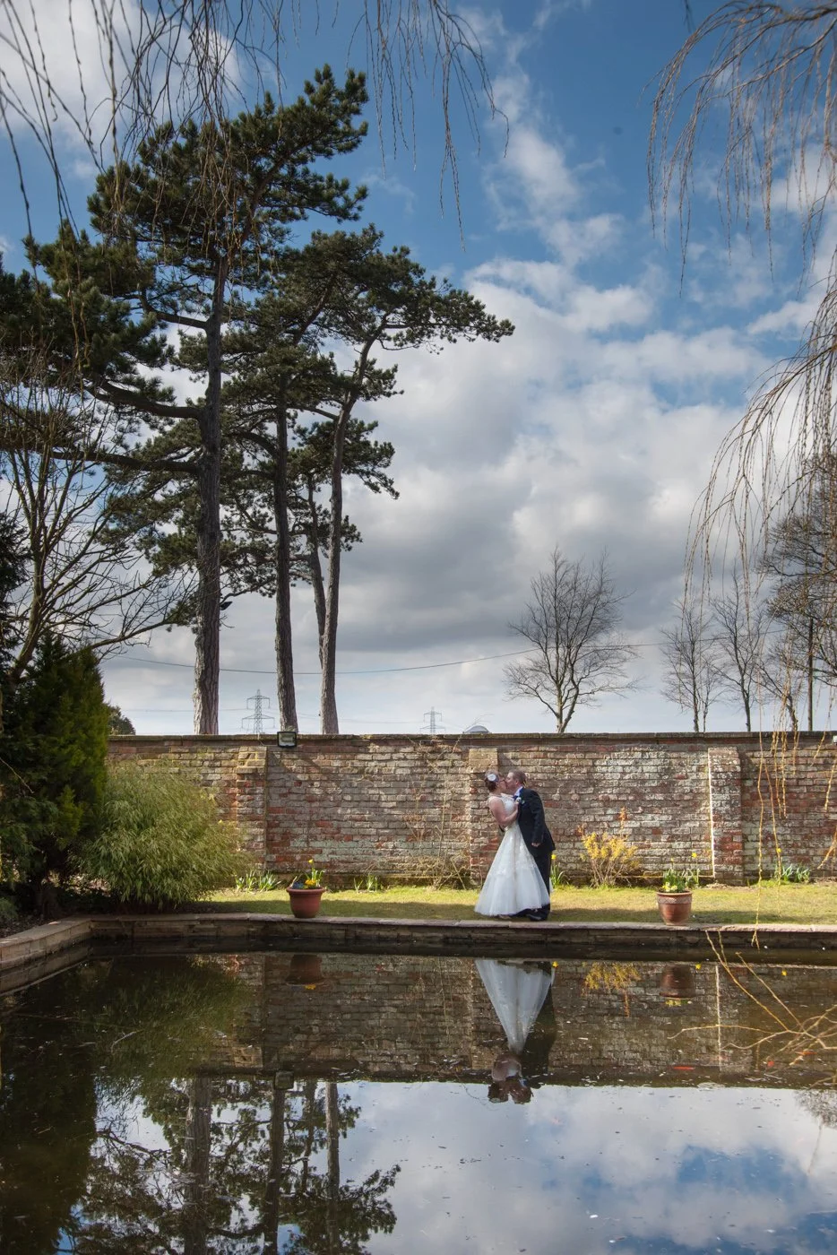 A bride and groom standing close together, kissing, in a garden area with a brick wall behind them, a small pond reflecting their image, and tall trees in the background under a cloudy sky.