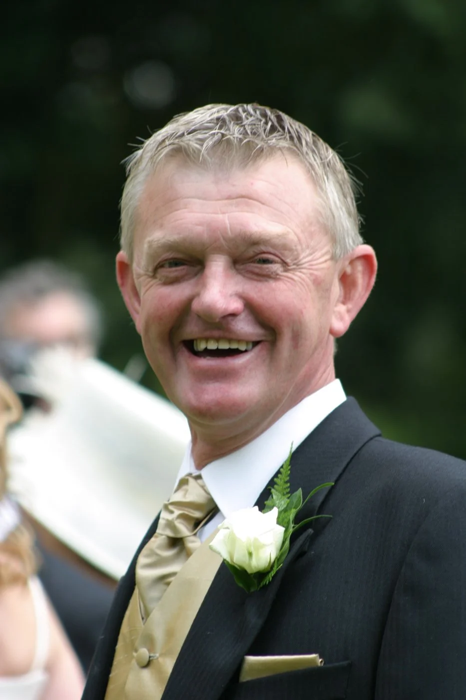 A middle-aged man in a tuxedo with a gold vest and tie, smiling outdoors on a wedding day, with a white boutonniere on his lapel.