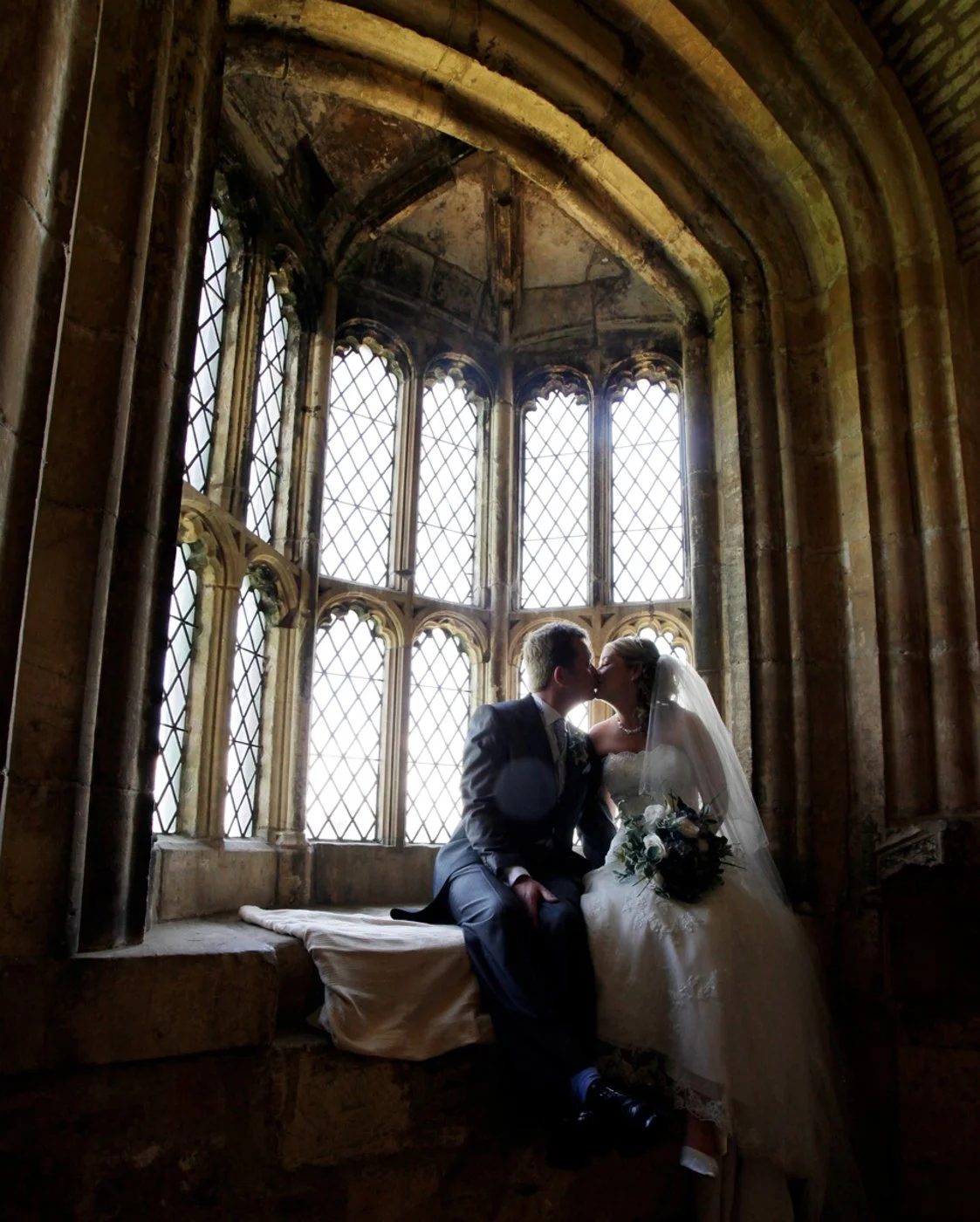 A bride and groom sharing a kiss inside a historic, Gothic-style stone building with large stained-glass windows.