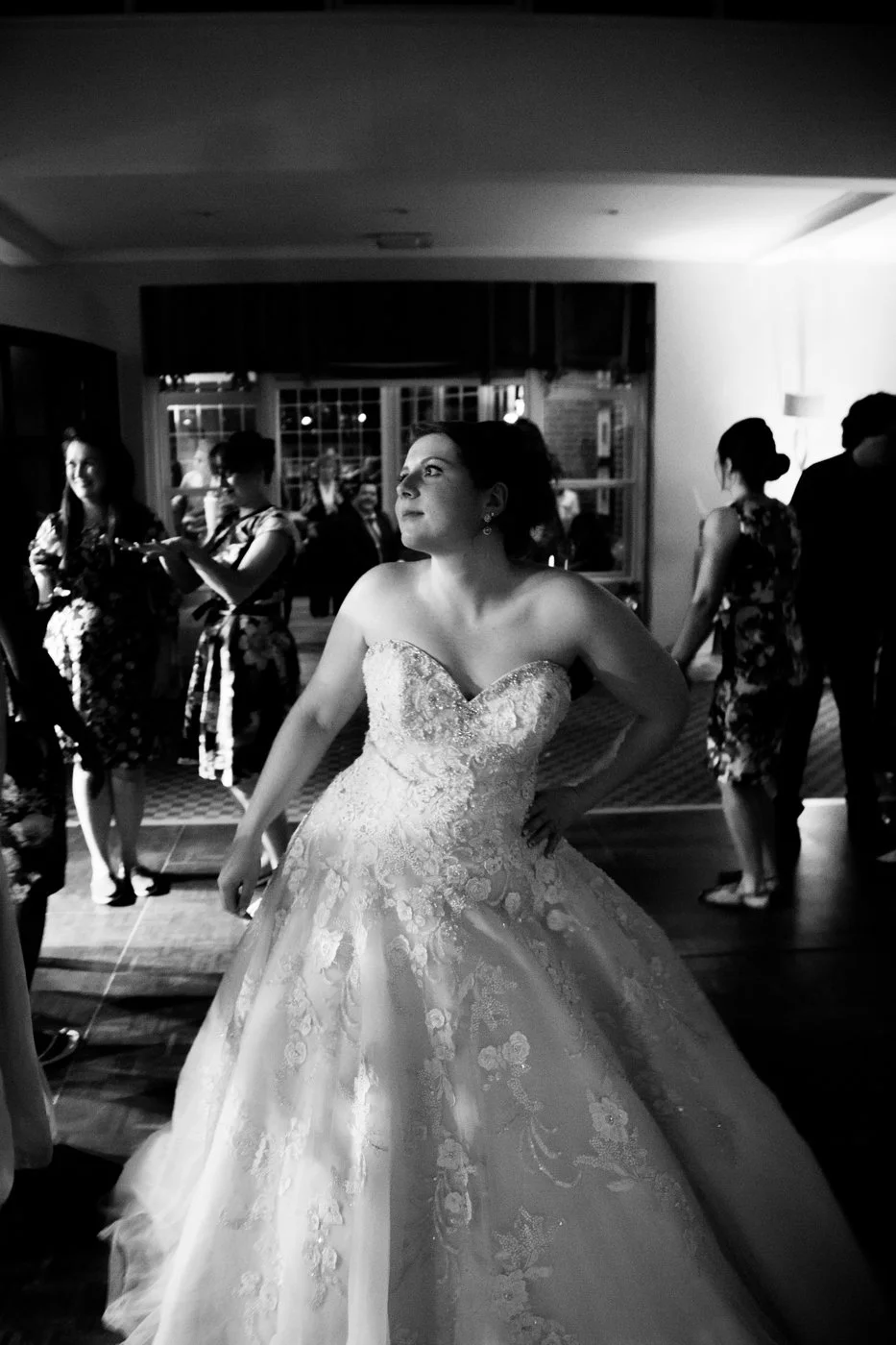 Black and white photo of a bride in a strapless wedding gown with lace embroidery, standing with her hand on her hip, surrounded by guests at a reception.