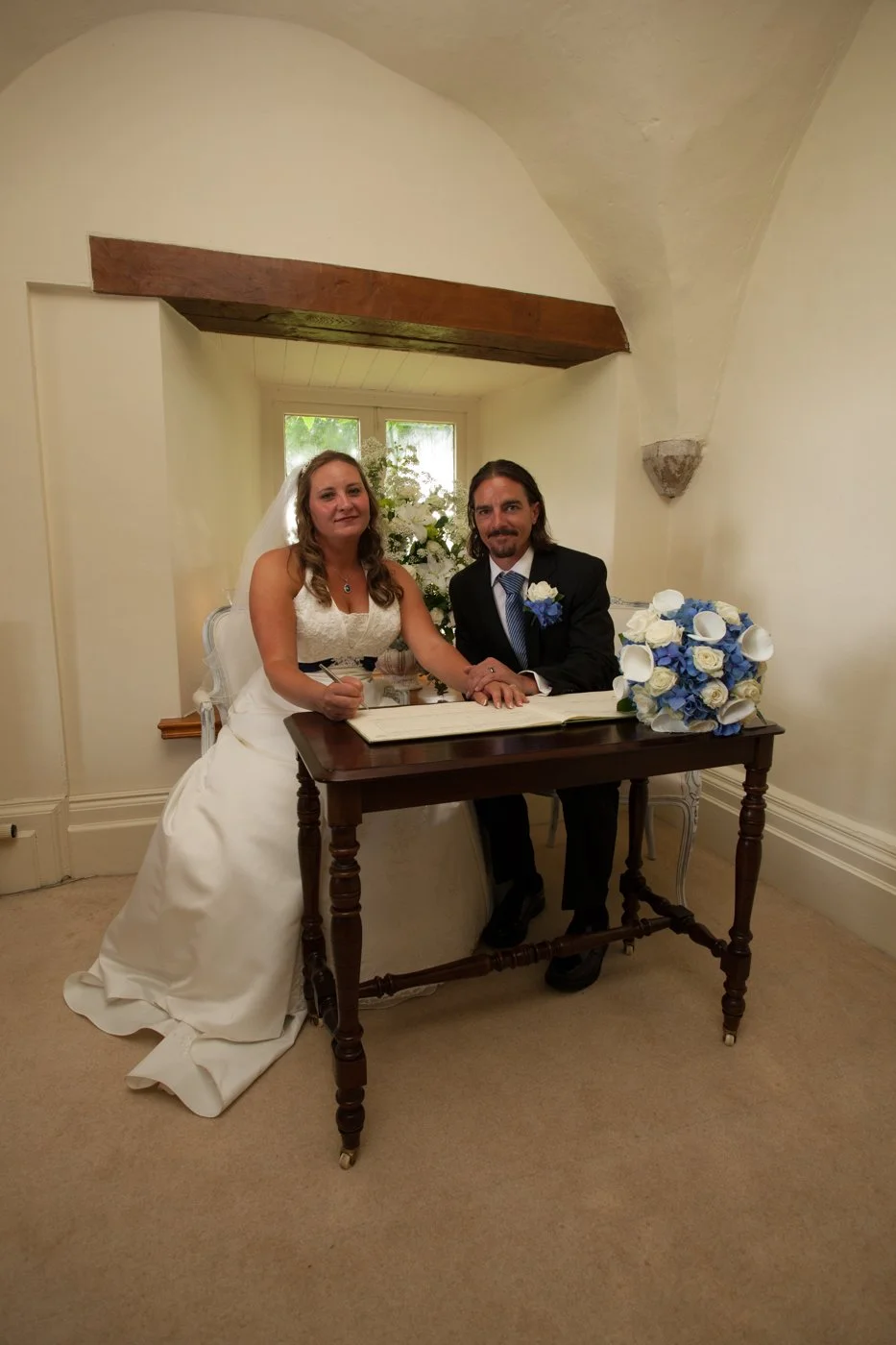 Bride and groom sitting at a wooden table during their wedding ceremony, signing documents, with a bouquet of white and blue roses on the table and a floral arrangement behind them.