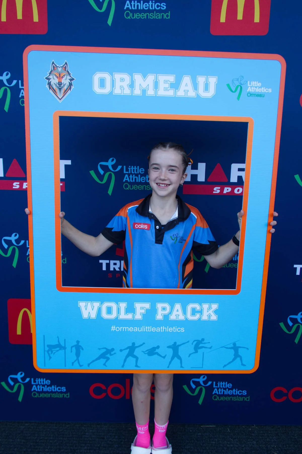 Smiling young girl in athletic uniform holding a large frame at an athletics event, with logos and background text indicating affiliation with Little Athletics Queensland and the Wolf Pack team.