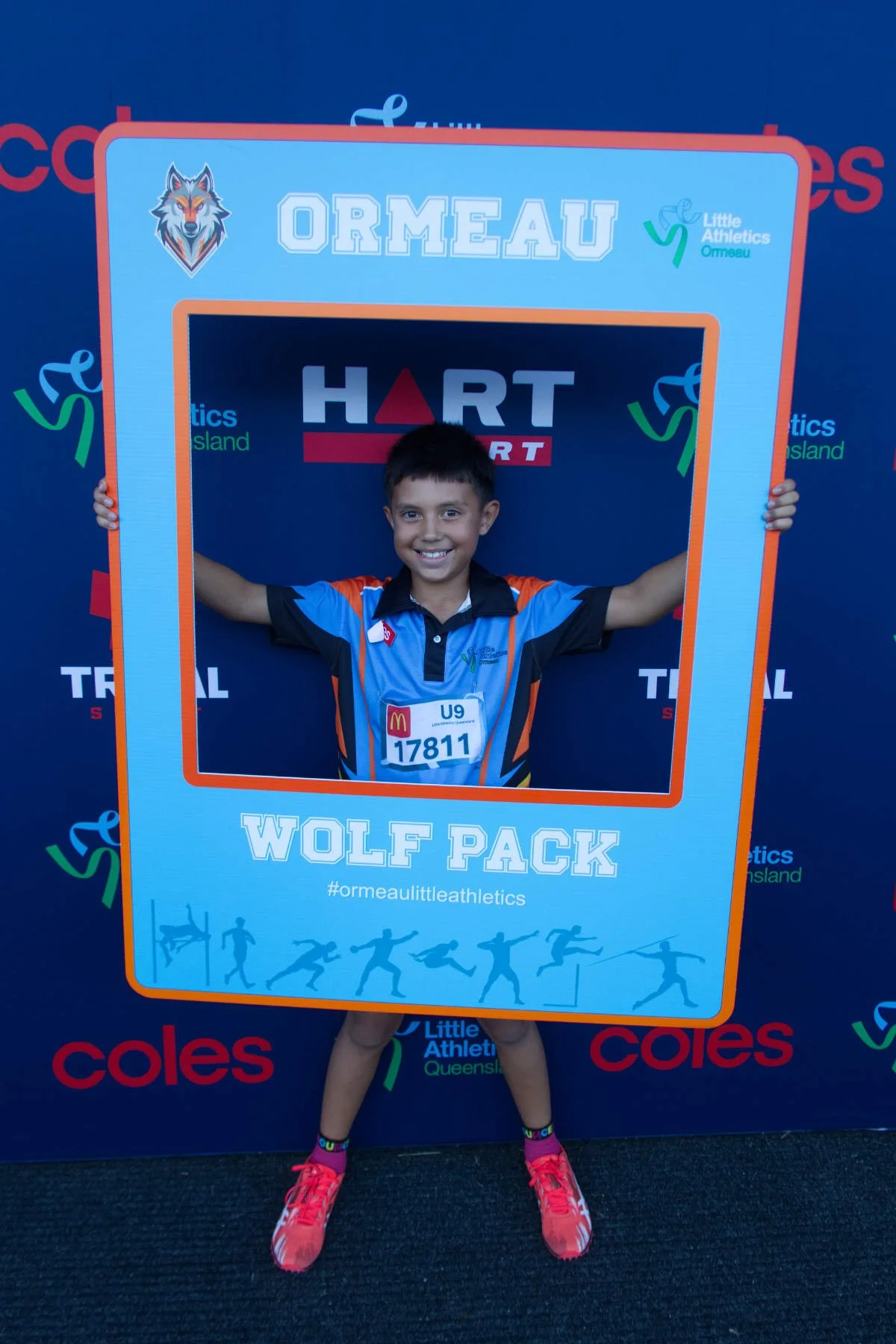 A young boy smiling while holding a large sports-themed frame at an athletic event. The frame features the words 'WOLF PACK' at the bottom and the hashtag '#ormeaulittleathletics,' with a wolf logo and a background of illustrated athletes. The boy is
