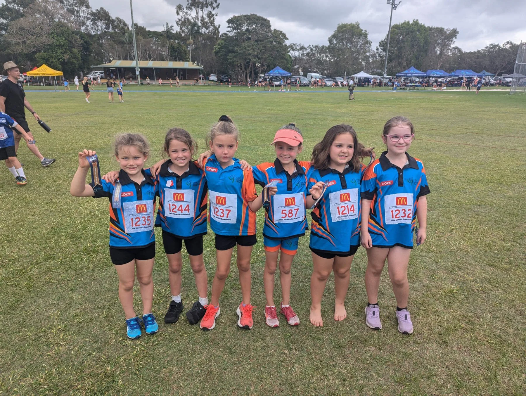 Group of six young girls wearing blue and orange sports uniforms standing on a grassy field, smiling and holding medals, with a track and spectators in the background.