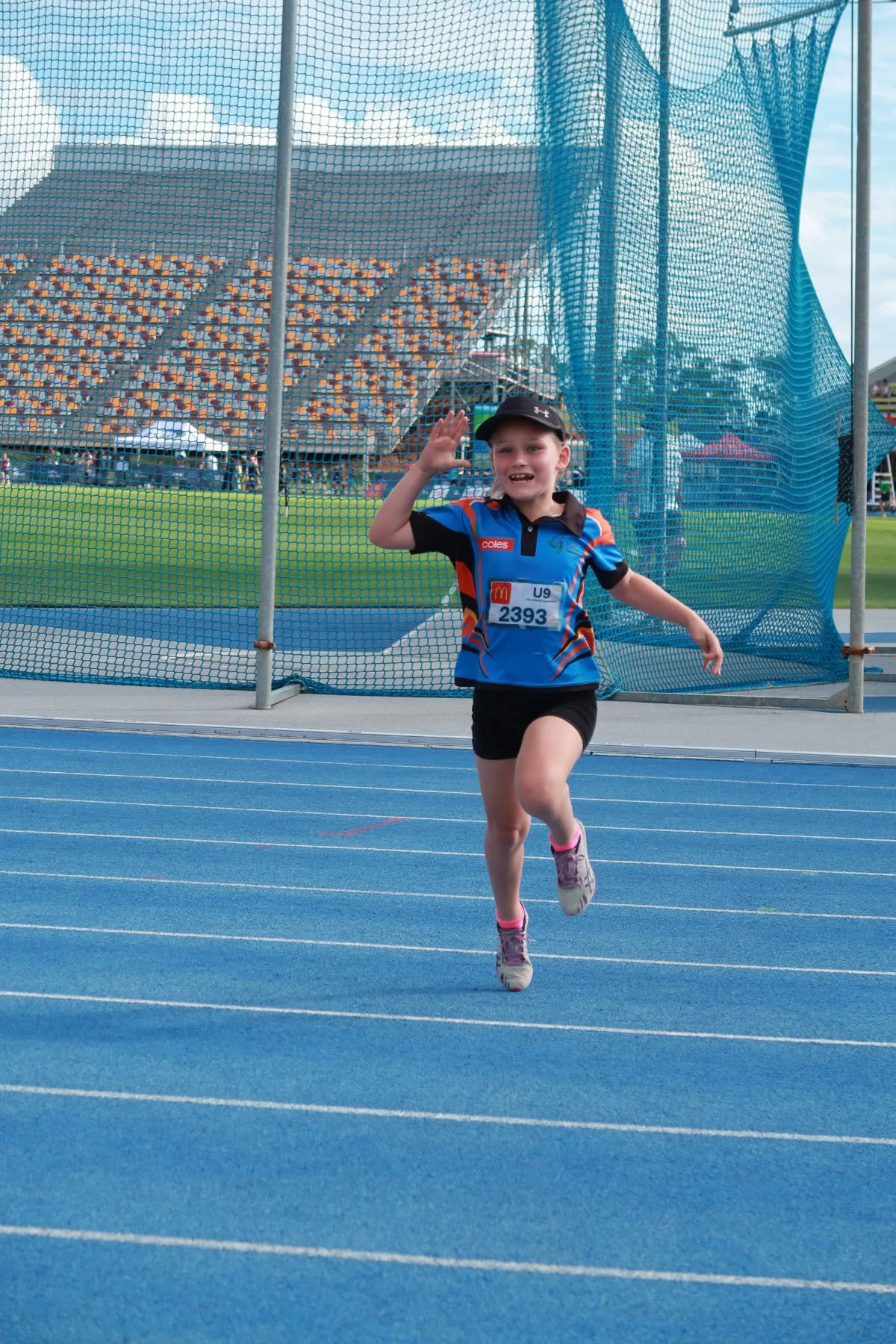 A young girl participating in a track race, running on a blue athletic track with a happy expression. She is wearing a blue sports shirt, black shorts, pink shoes, and a black cap, with race bib number 2393. In the background, a sports field, bleache