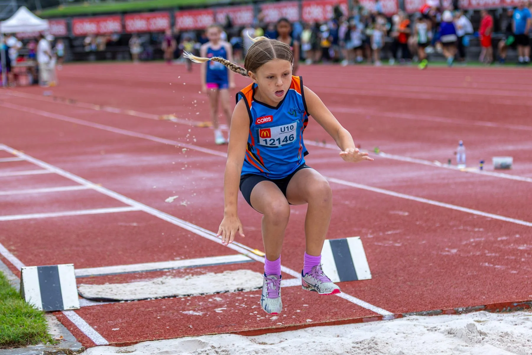 Young girl in athletic outfit jumping over a hurdle on a track field during a race, with other competitors and spectators in the background.