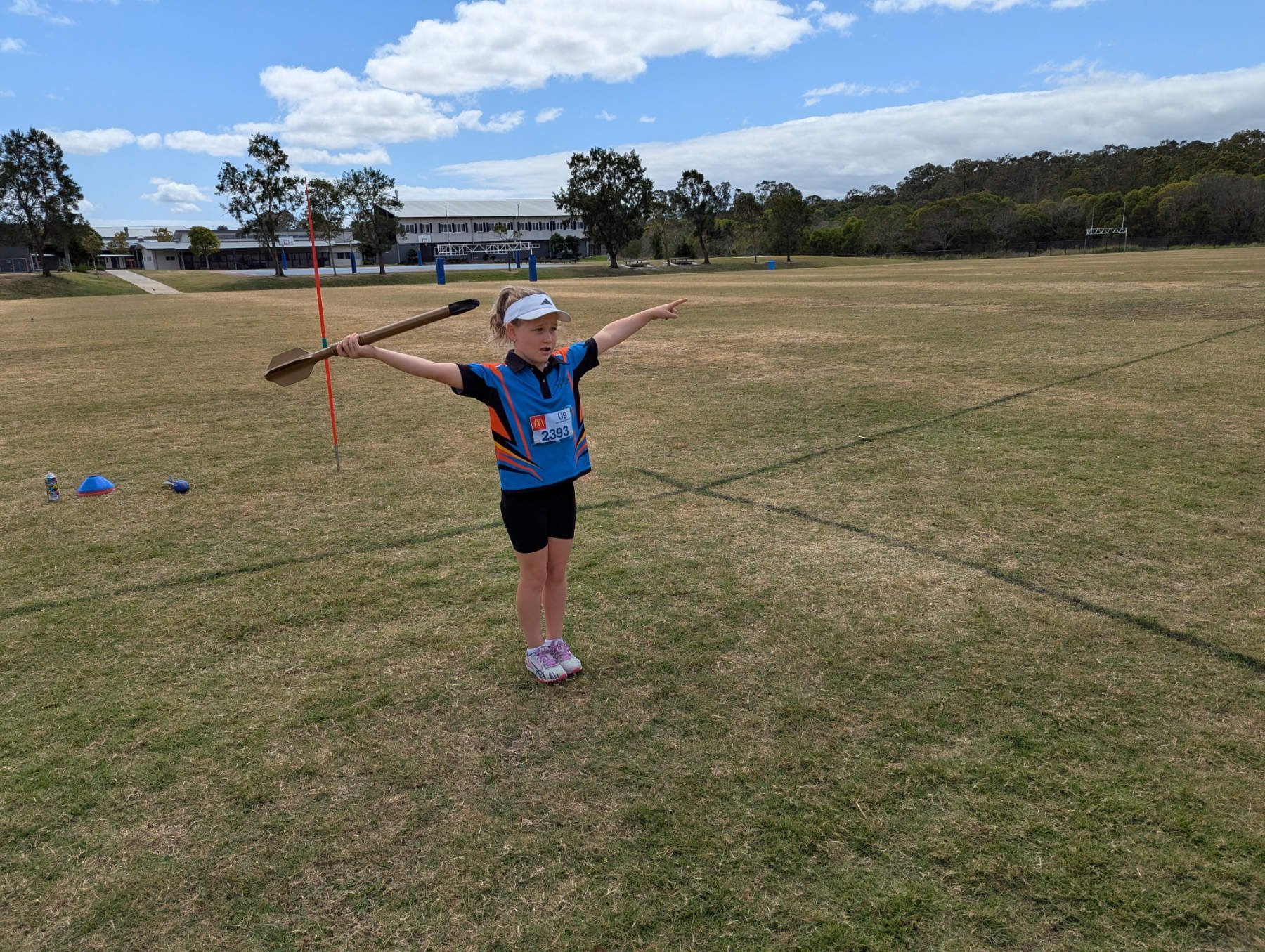 Young girl in athletic clothing standing on a sports field with a javelin in her right hand, pointing with her left arm, under a partly cloudy sky.