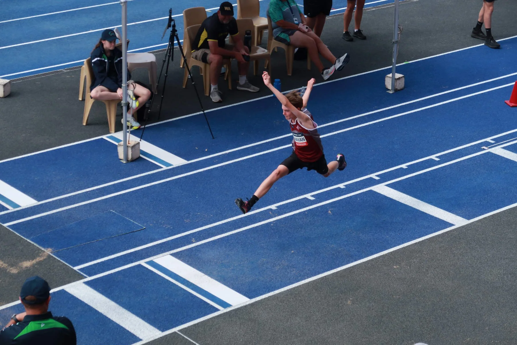 A young male athlete in a red uniform is mid-air, landing after a long jump on a blue track surface at a stadium, with officials and spectators watching.