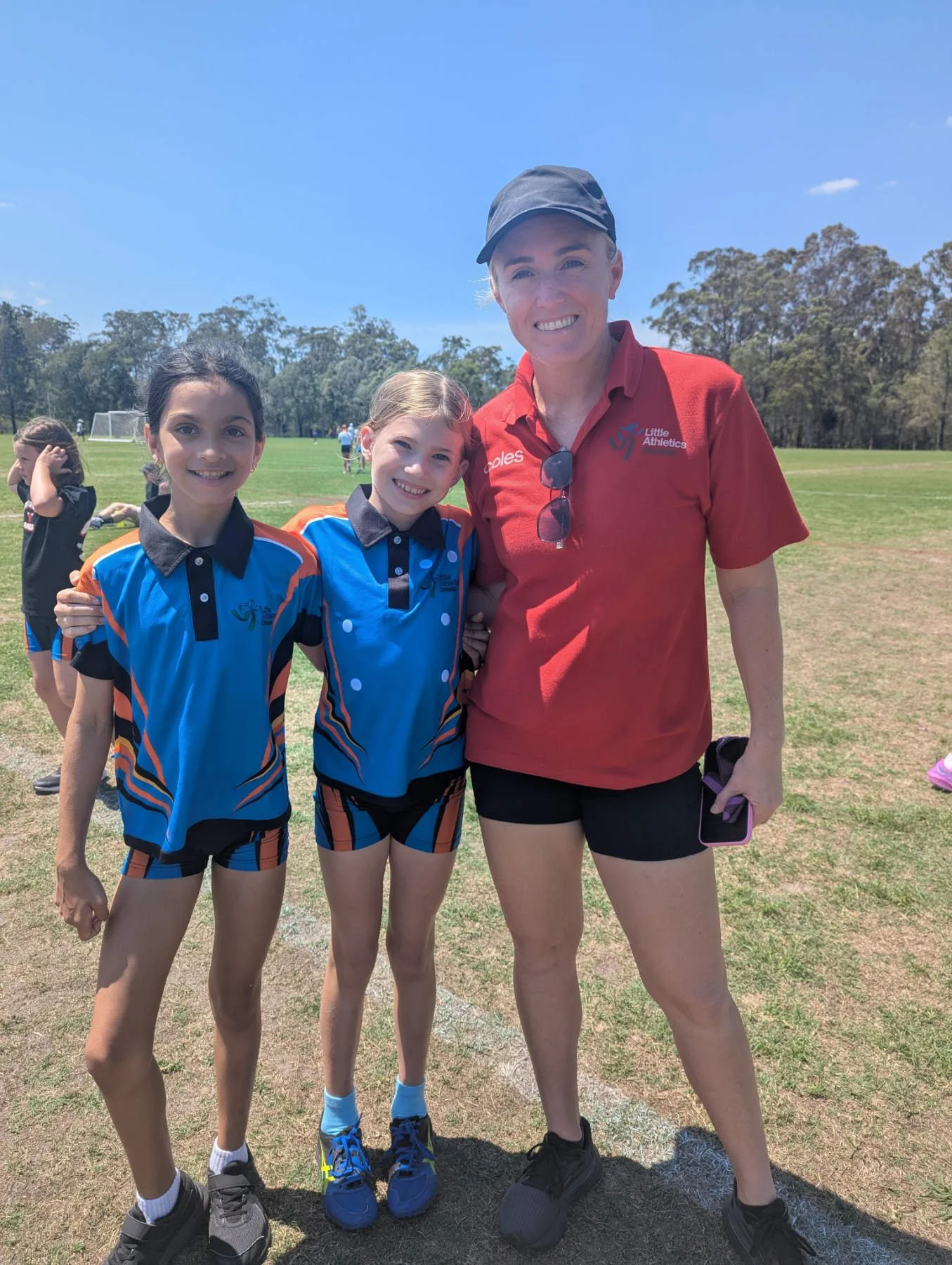 A woman with a cap and sunglasses around her neck standing with two young girls in sports uniforms on a field, smiling at the camera, with other children in the background.