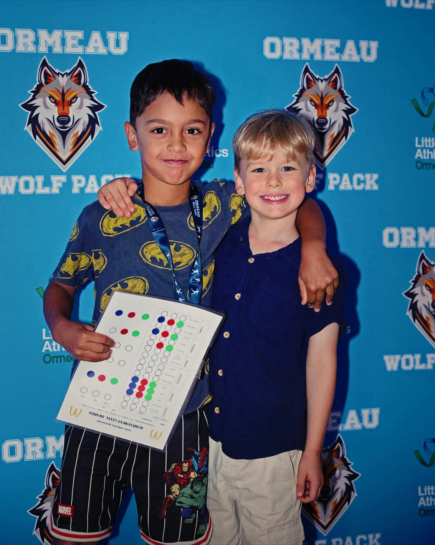 Two young boys standing side by side, smiling, at an event with a blue backdrop that has logos and text. One boy has dark hair, wearing a Batman shirt and striped shorts, holding a sheet of paper with colorful dots. The other boy has blonde hair, wea