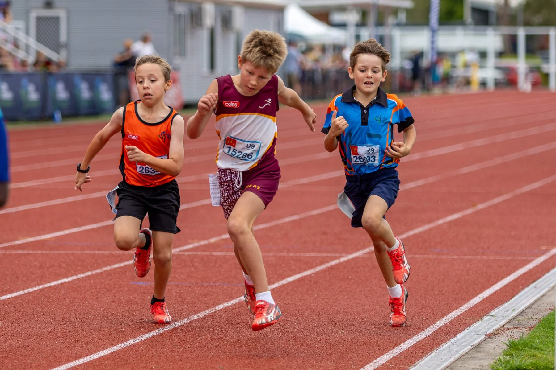 Three young boys running a race on a red outdoor athletic track, wearing colorful athletic uniforms and red running shoes, with an overcast sky and spectators in the background.