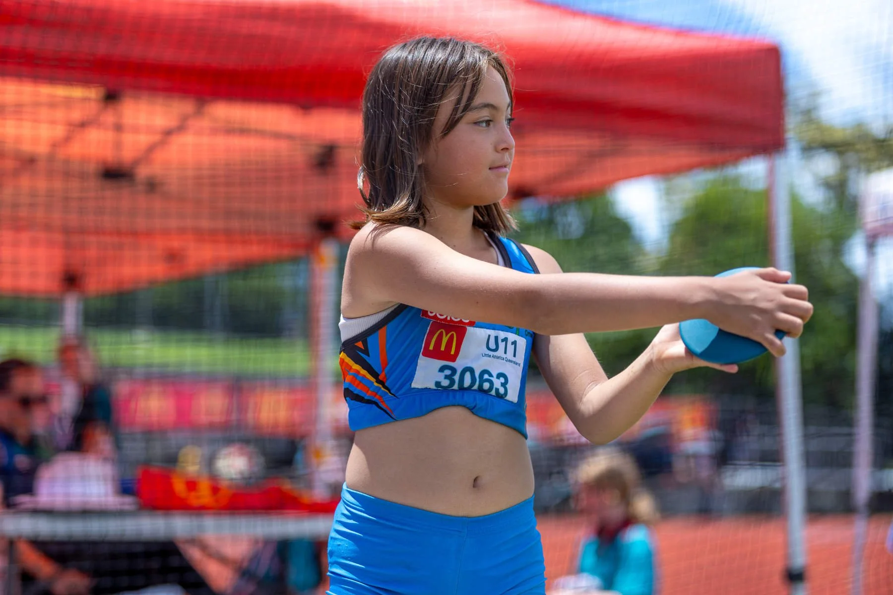 Young girl in athletic clothing holding a shot put during a track and field event, with spectators and orange barriers in the background.