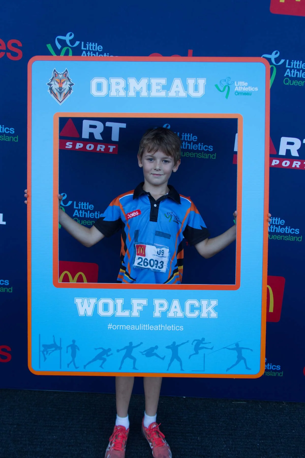 Young boy holding a large framed sign at a Little Athletics event. The sign has a wolf logo, the name 'Ormeau,' and mentions 'Wolf Pack.' The boy is wearing a sports uniform with sponsor logos, a race bib number 26093, and red sneakers.